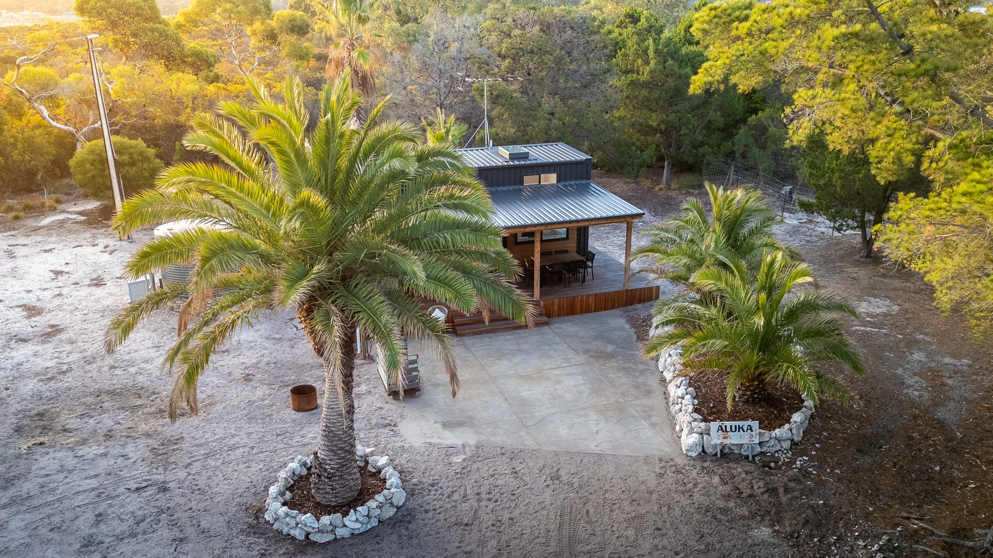 A small house with a metal roof and porch, surrounded by palm trees and desert landscape, with a sign that reads 'ALUKA' in front.