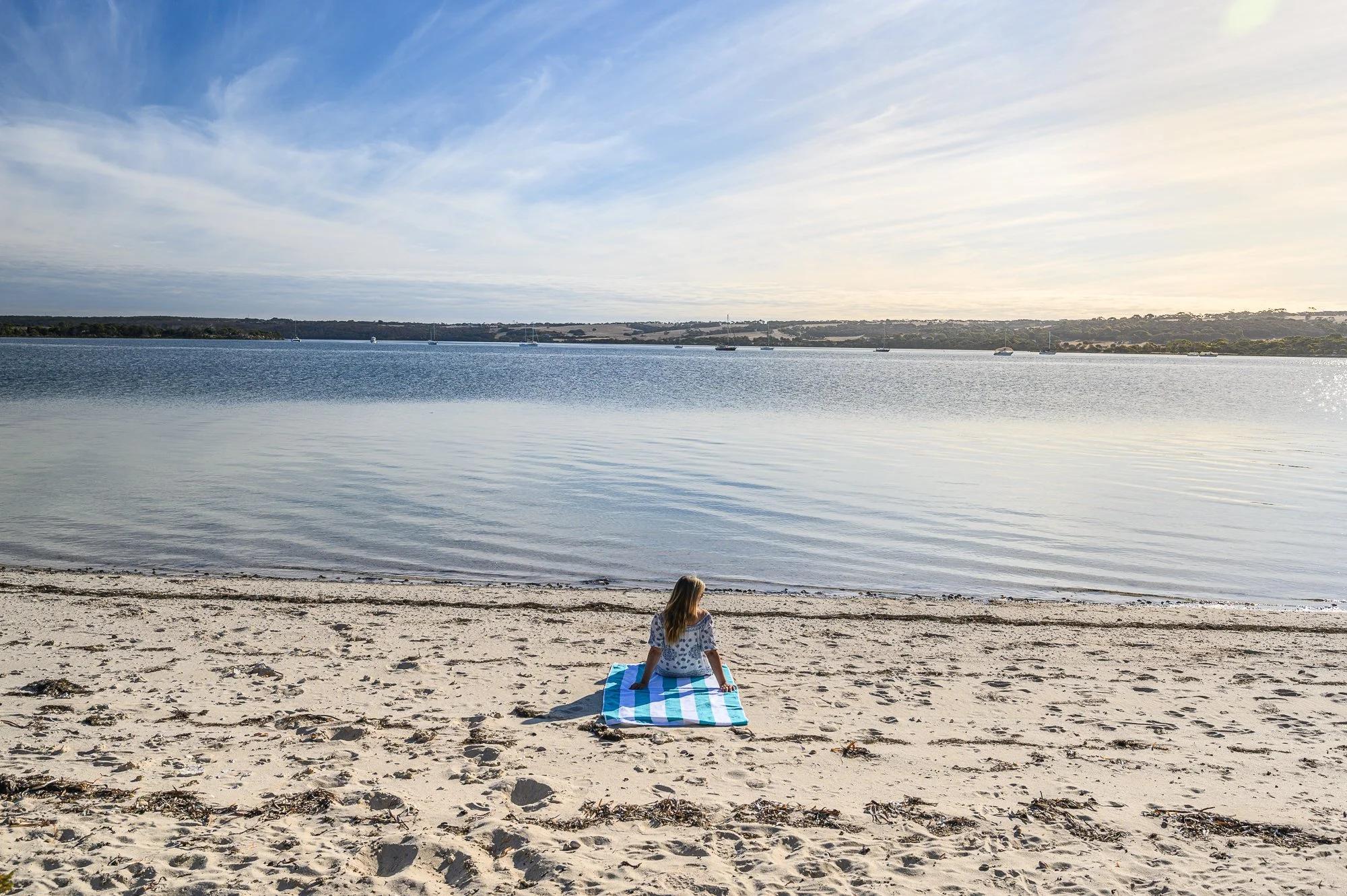 A girl sitting on a striped towel on a sandy beach, looking at the calm water and distant sailboats under a partly cloudy sky.