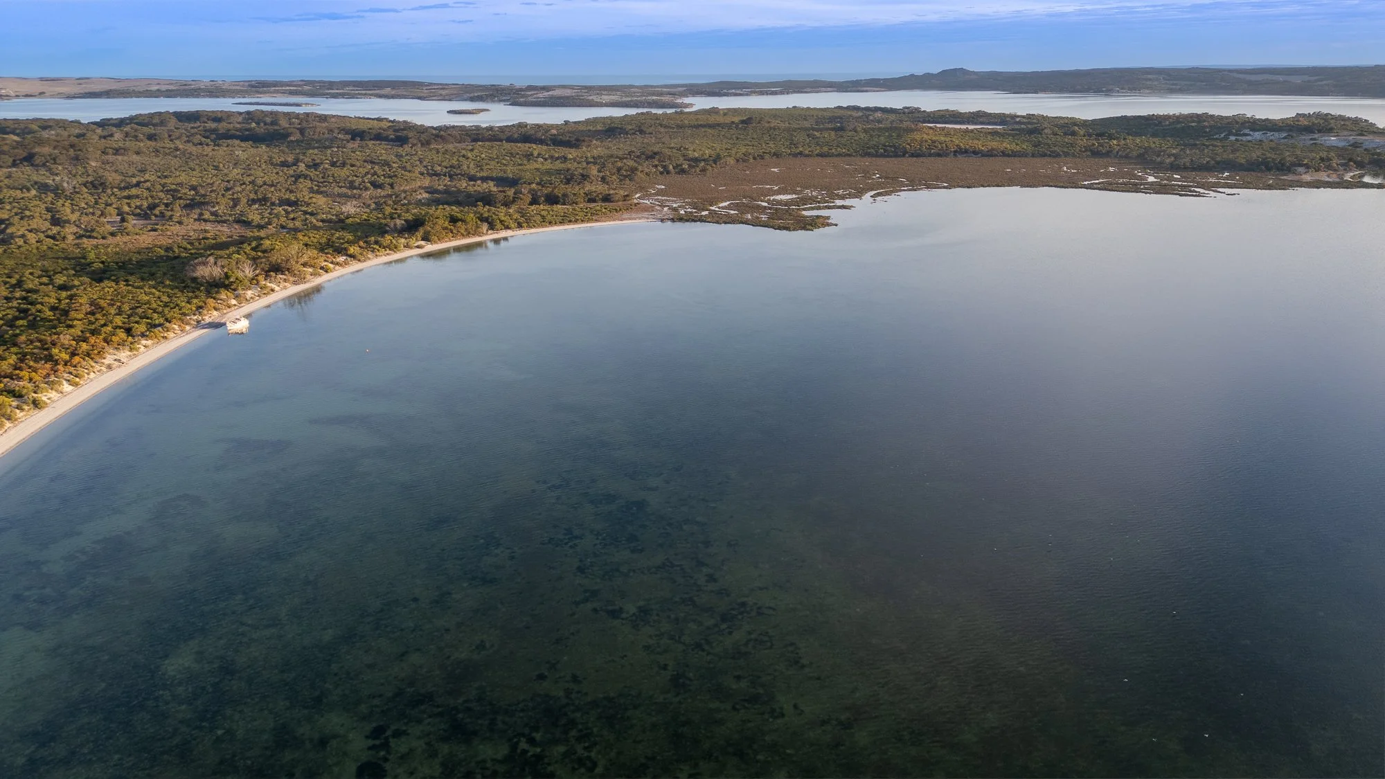 Aerial view of a large bay with clear water, a sandy shoreline, and dense forested land in the background, under a partly cloudy sky.
