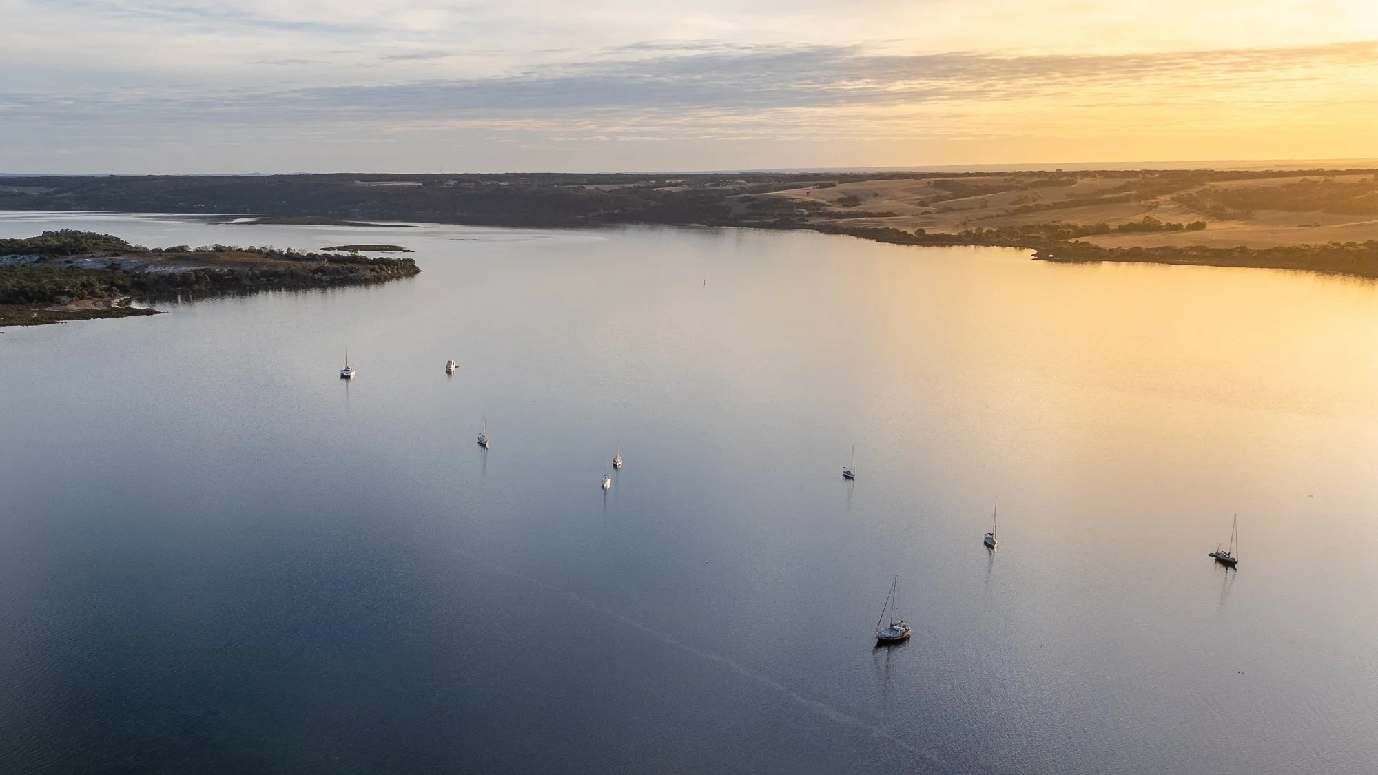 Aerial view of a calm body of water with several anchored sailboats, surrounding land with trees and open fields, under a partly cloudy sky with a warm sunset glow.