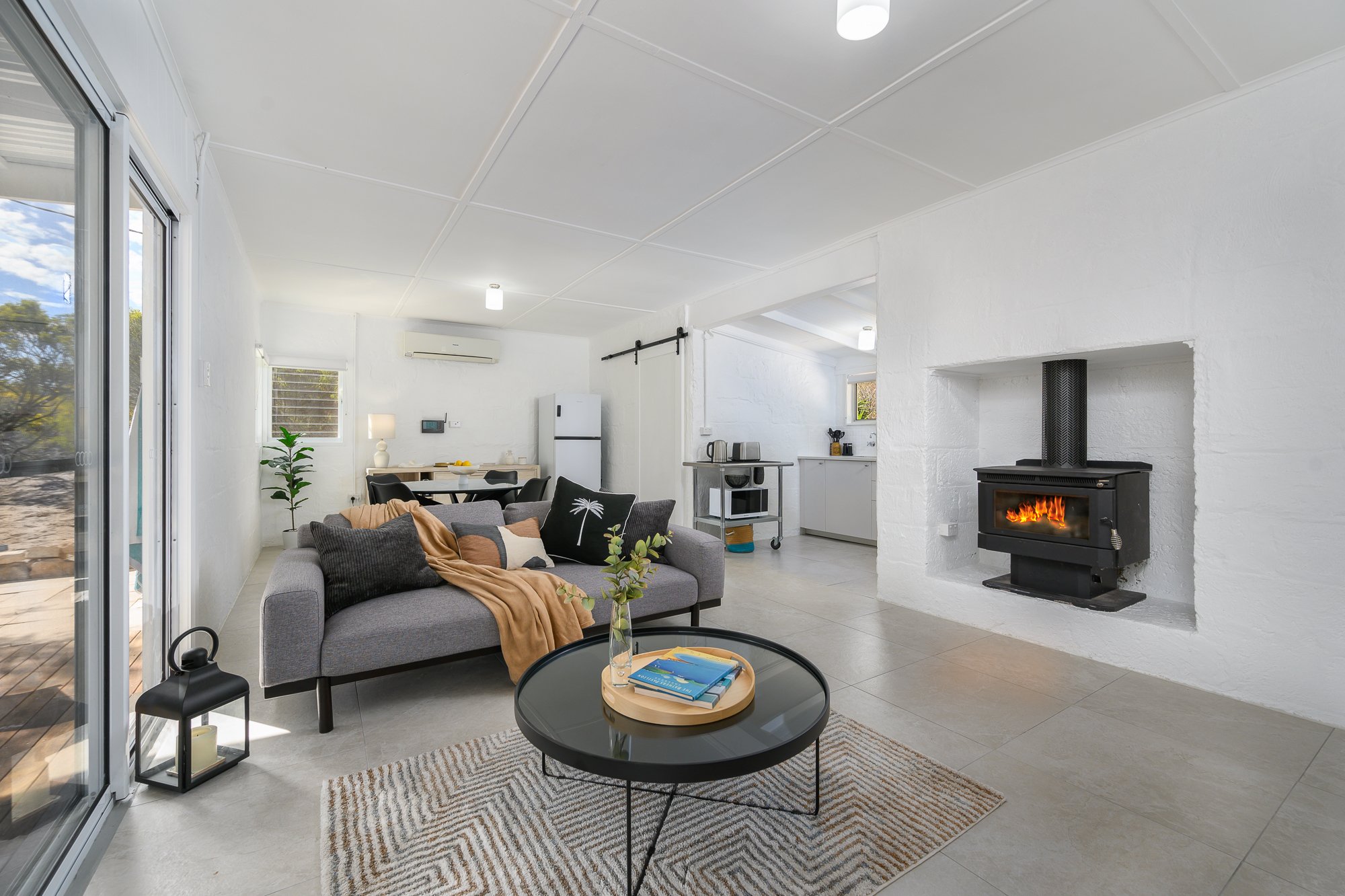 Living room with a gray sofa, decorative pillows, a throw blanket, a round black coffee table with books and a vase, a fireplace with a fire, a dining area with a table and chairs, a kitchen area with white cabinets, and sliding glass doors leading o