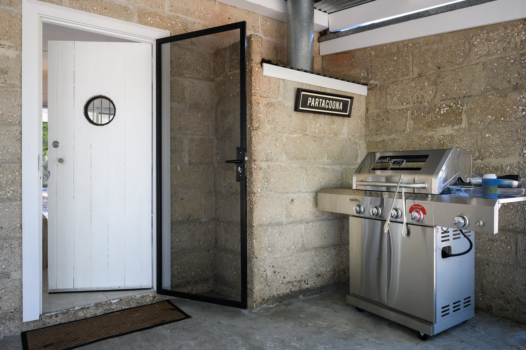 A stainless steel outdoor barbecue grill next to a brick wall, with a black and white sign that reads 'PARTACOONA' and a white wooden door with a small round window.