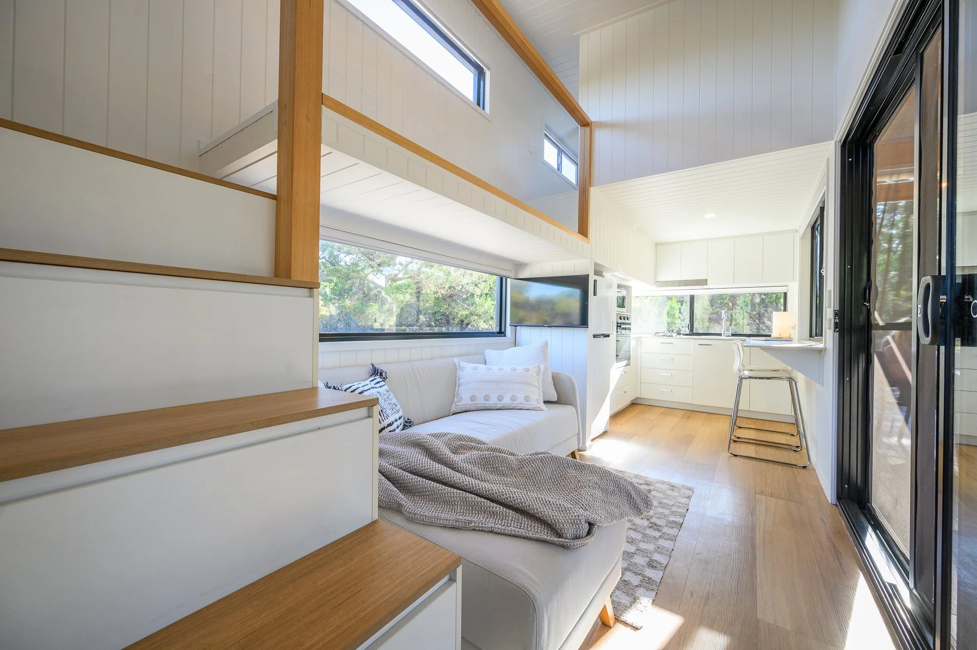 Interior of a modern, bright living space with white walls and wooden accents, featuring a white couch with cushions, a large window, and a kitchen area with white cabinets and a barstool by a counter.