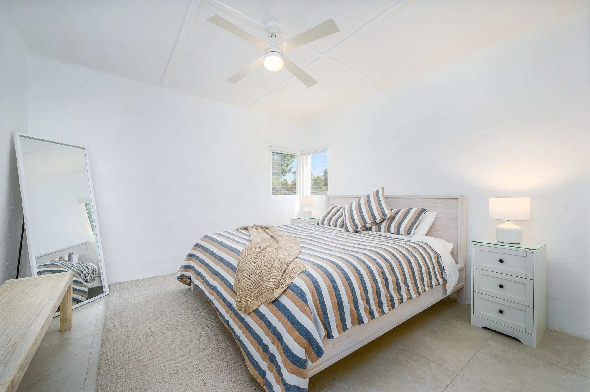 A bedroom with a white bed frame and striped bedding, two pillows, a beige throw blanket, a white bedside table with a lamp, a window, a ceiling fan, a mirror leaning against the wall, a bench, and beige tiled floor with a rug.