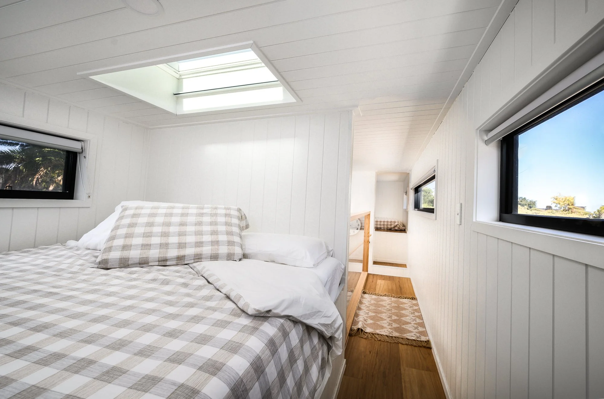 A small bedroom with white paneled walls and ceiling, a skylight, and small horizontal windows. A bed with plaid bedding and pillows is on the left, with the head of the bed near the window. A small wooden side table is at the foot of the bed, with a