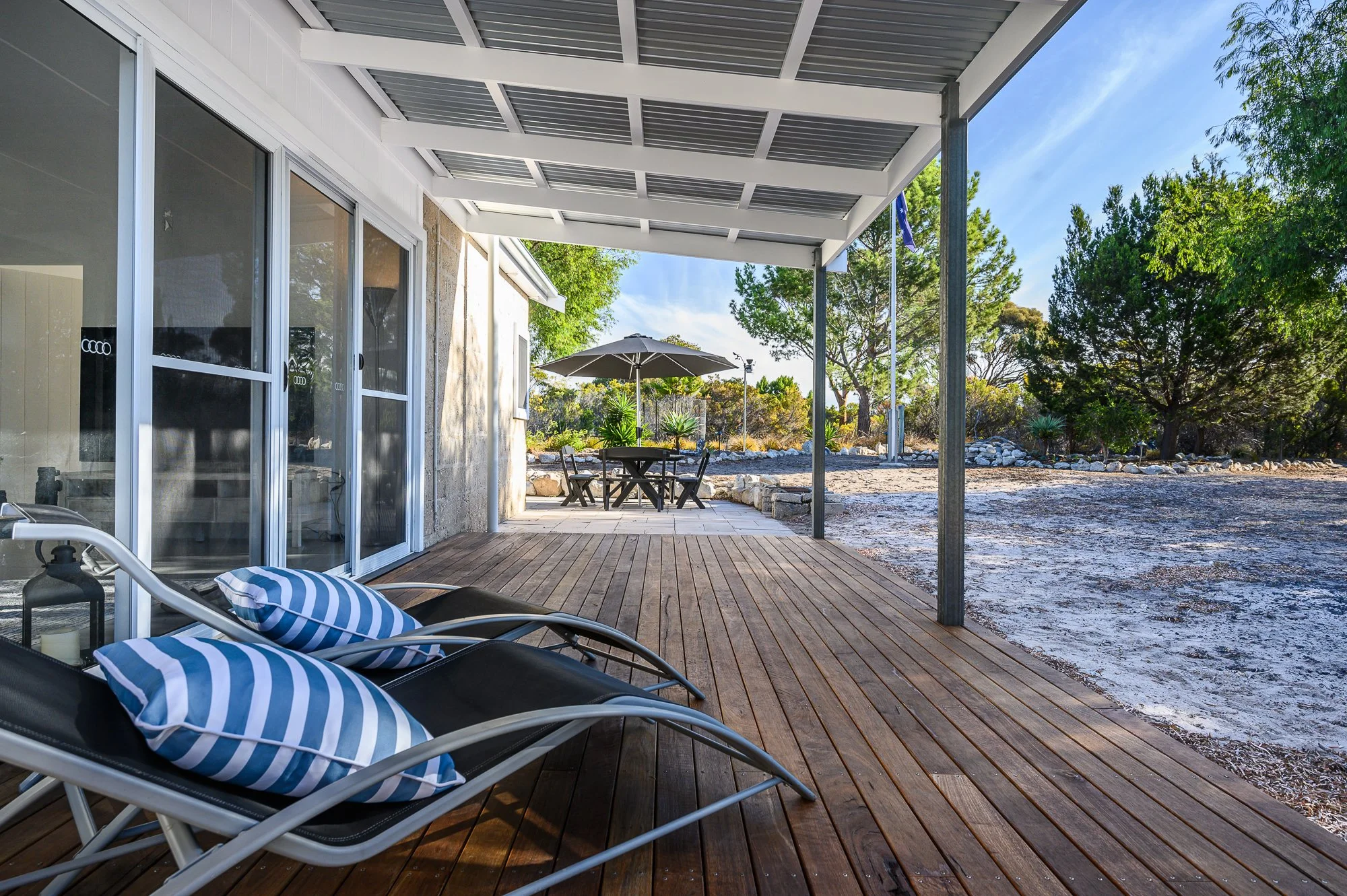 A covered wooden patio with two lounge chairs with striped pillows in the foreground, a round table with an umbrella and chairs in the background, surrounded by trees and open outdoor space.