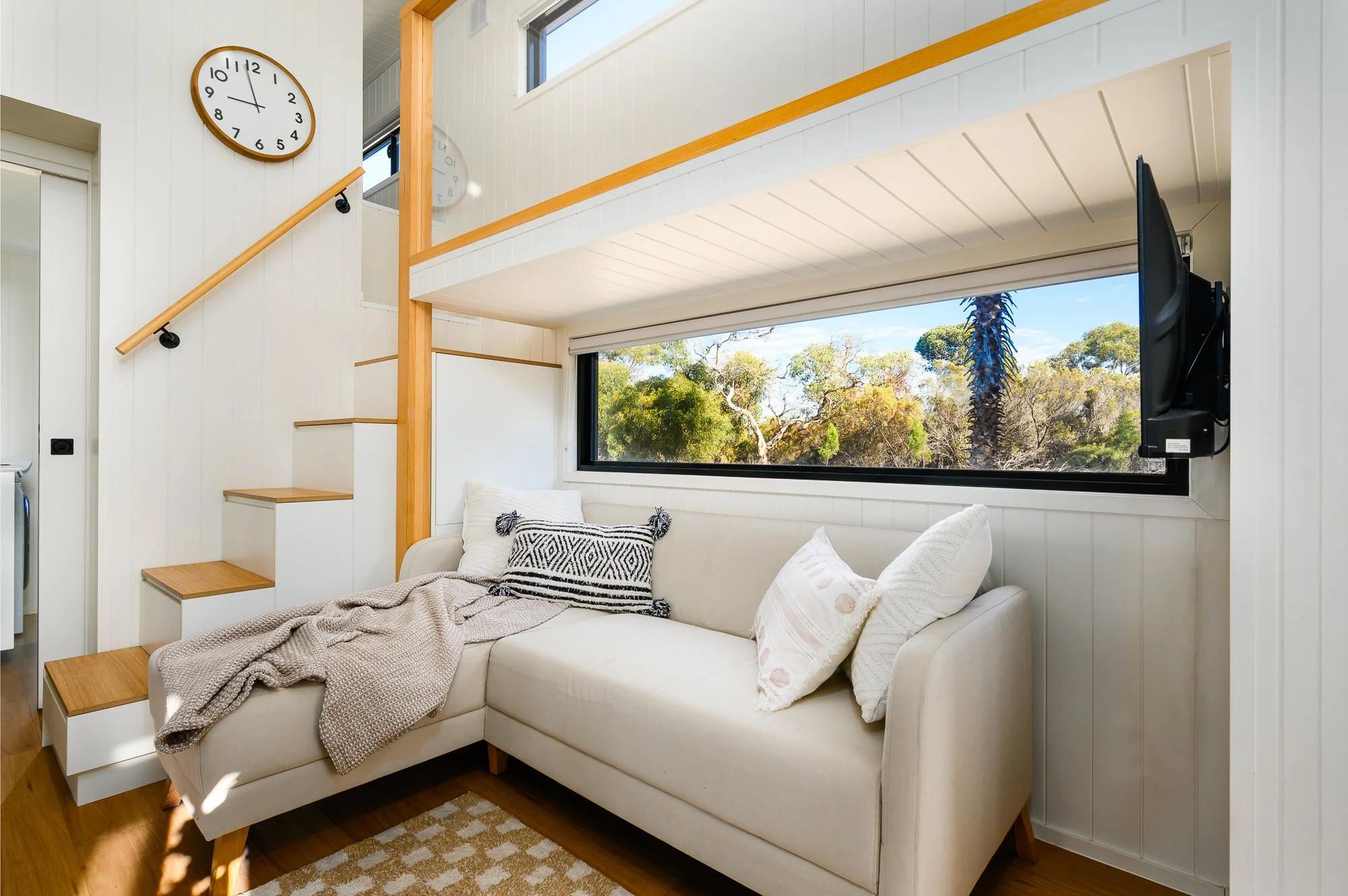 Living room with a white sofa, a large horizontal window with a view of trees, a wall clock, and a staircase with wooden steps and a black railing.