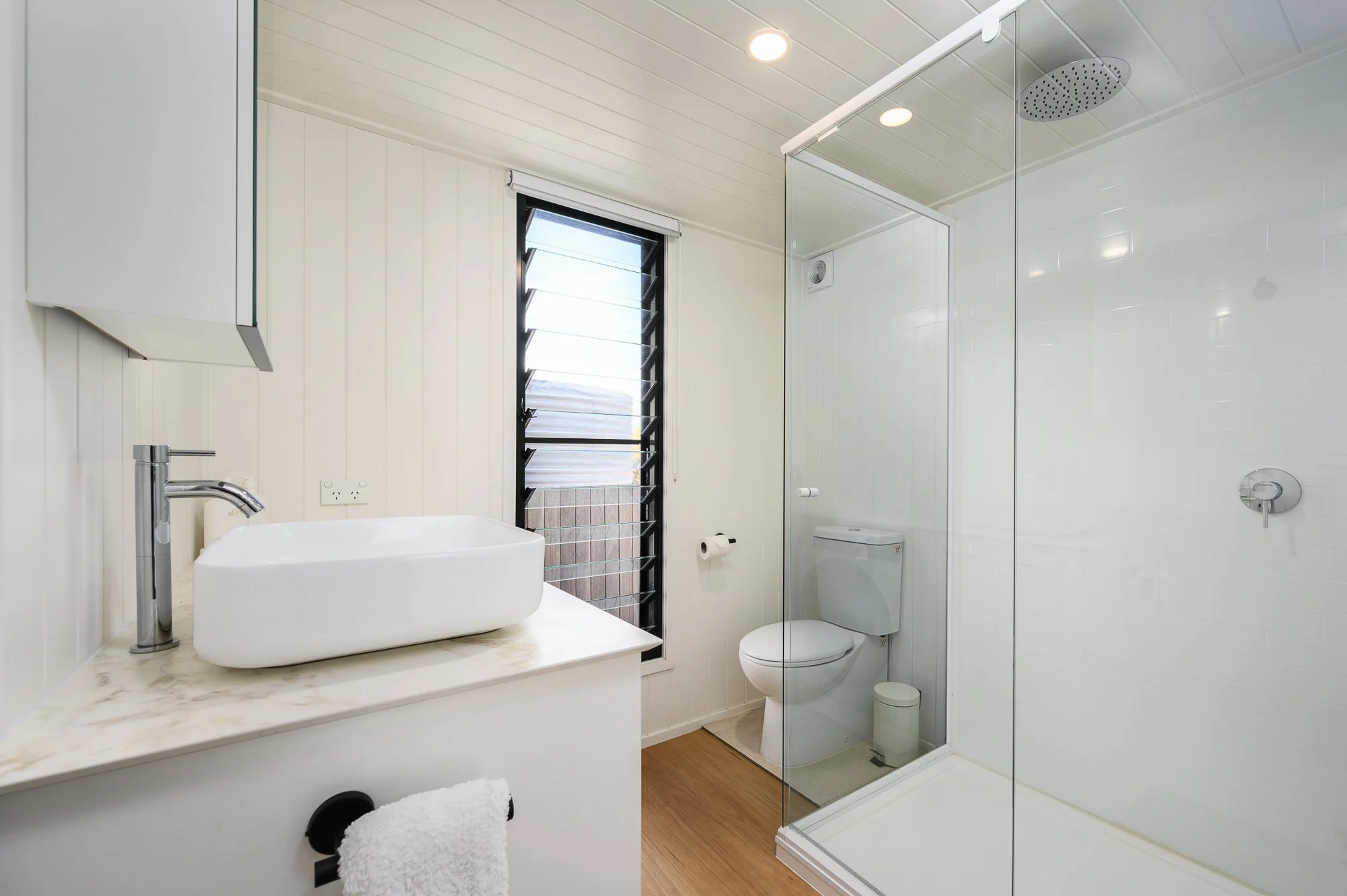 Modern bathroom with white walls, wooden flooring, a glass-enclosed shower with a rainfall showerhead, a toilet, a window with black horizontal slats, and a white vessel sink on a marble countertop.