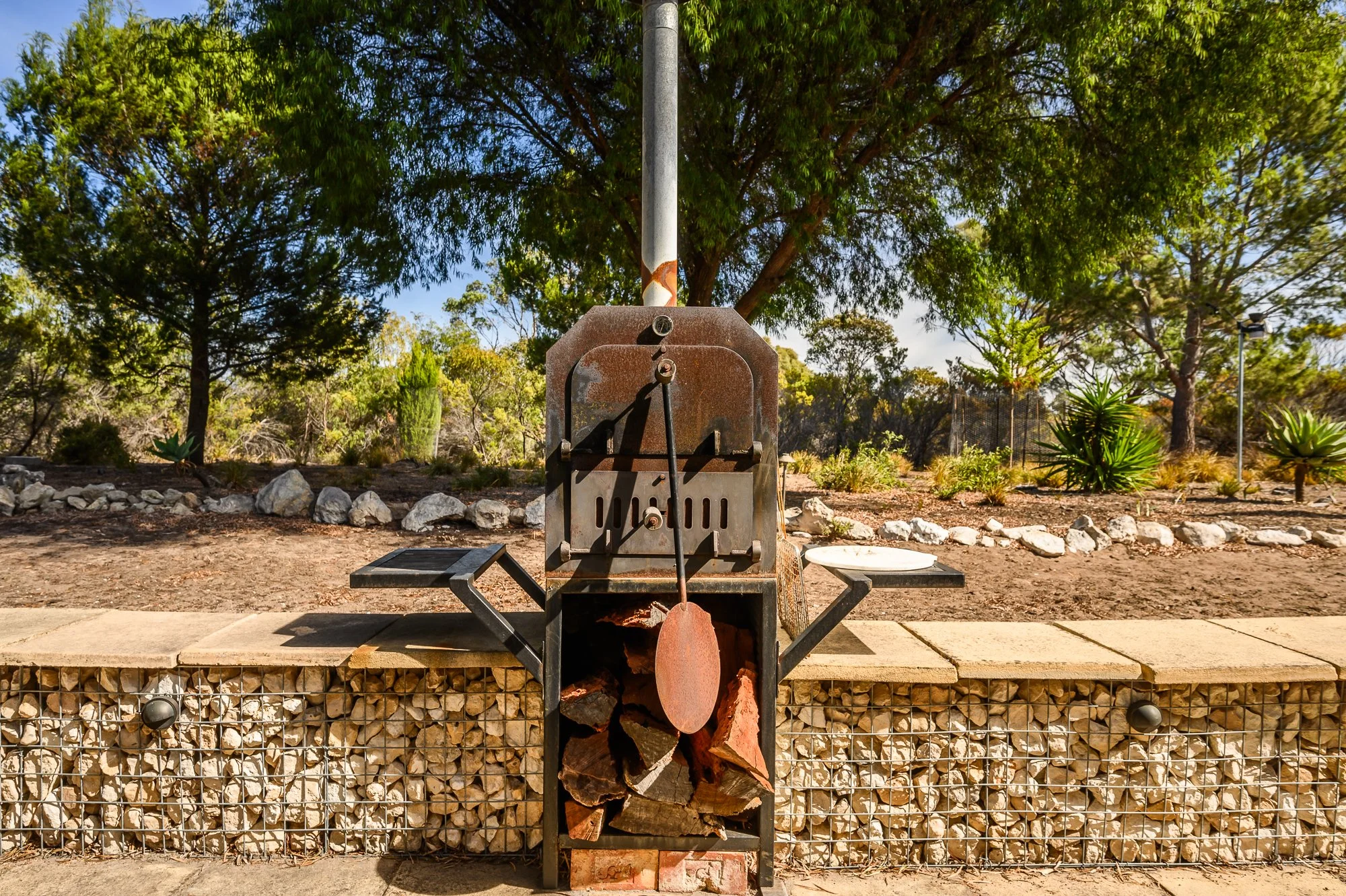 Rusty outdoor barbecue grill with cooking tools, including a spatula, attached to the front, positioned on a stone wall with firewood inside. The background features a landscaped garden with bushes and large trees under a clear sky.