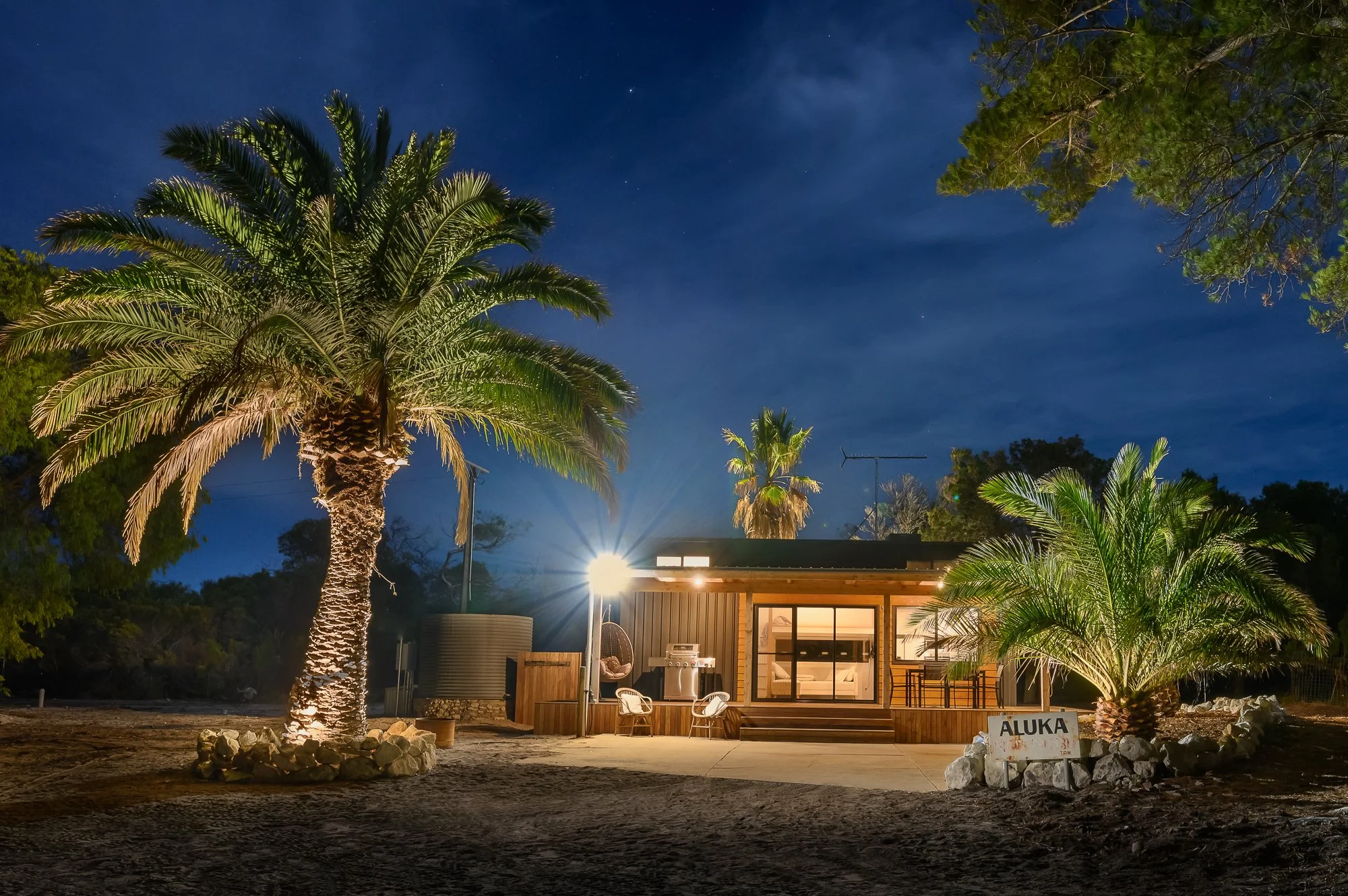 Nighttime view of a small wooden house with large glass sliding doors, illuminated from inside, surrounded by palm trees, with a sign reading 'ALUKA' near one of the trees, and a bright outdoor light illuminating the scene.