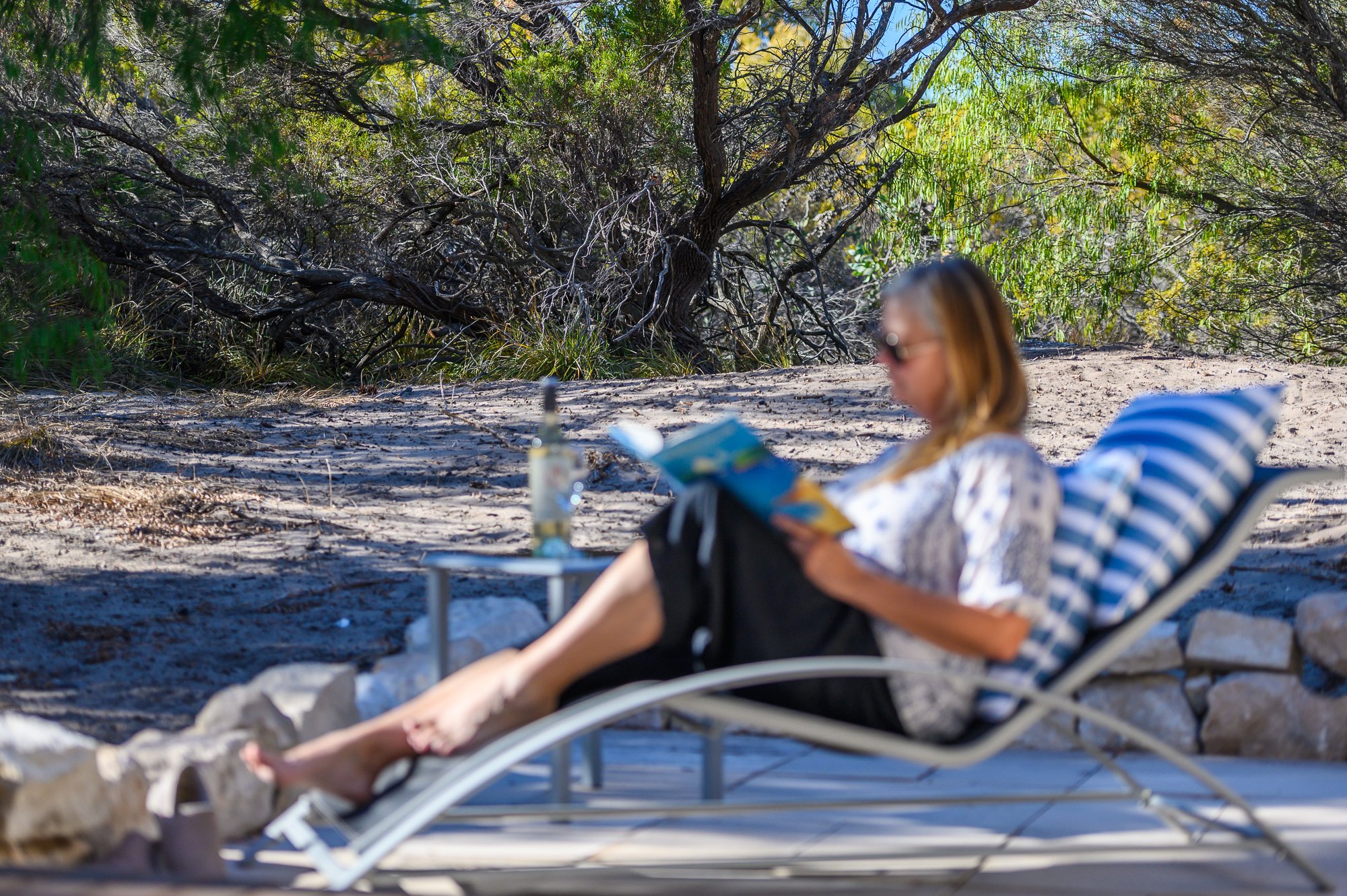 A woman relaxing on a lounge chair, reading a book near a sandy area with trees in the background.