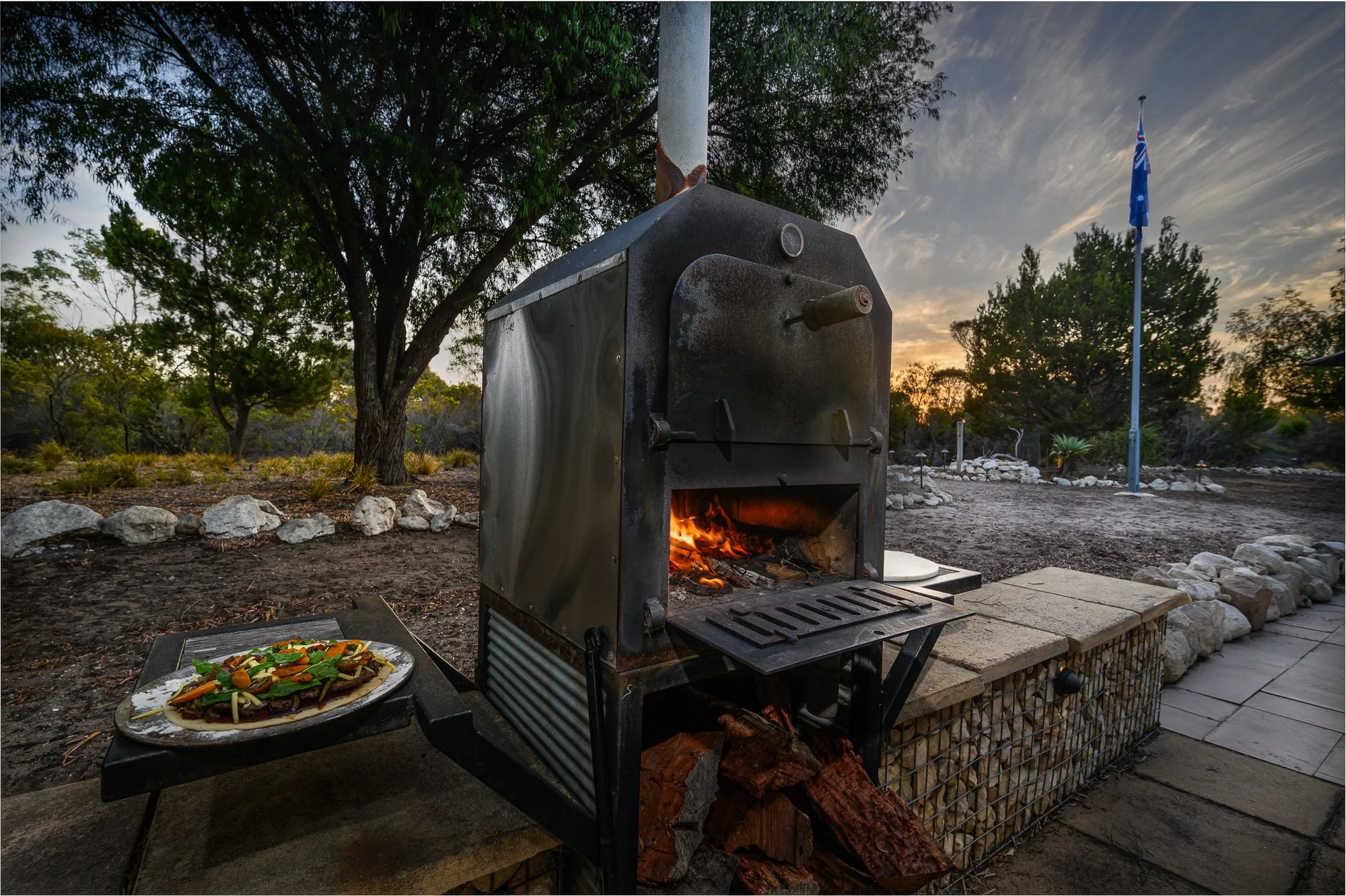 A wood-fired pizza oven with a flame inside, positioned outdoors during sunset, with a plate of assorted pizza toppings on a nearby table and a flagpole with an Australian flag in the background.