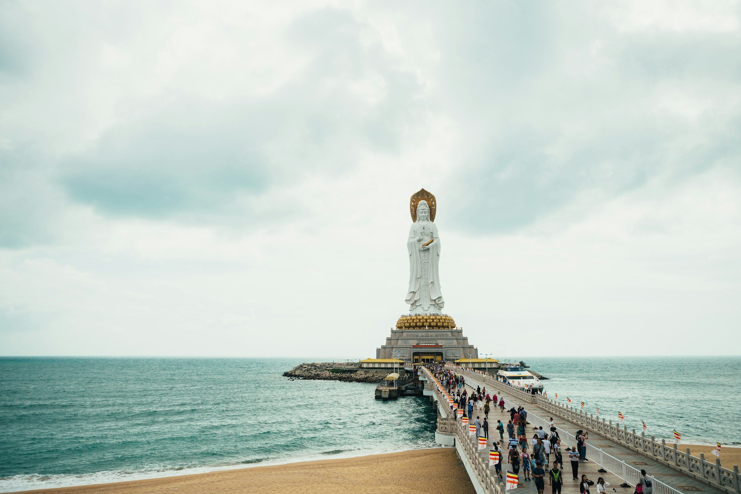 Large white statue of a deity on a platform at the end of a pier, with tourists walking towards it over a bridge, ocean on both sides, cloudy sky overhead.