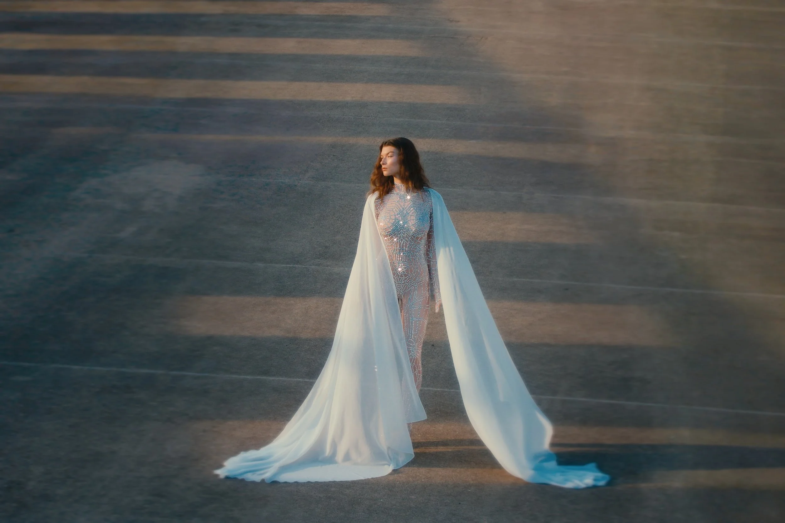 Woman in a sparkling dress with long flowing fabric, standing on an open concrete surface, with shadows and sunlight creating a dramatic effect.