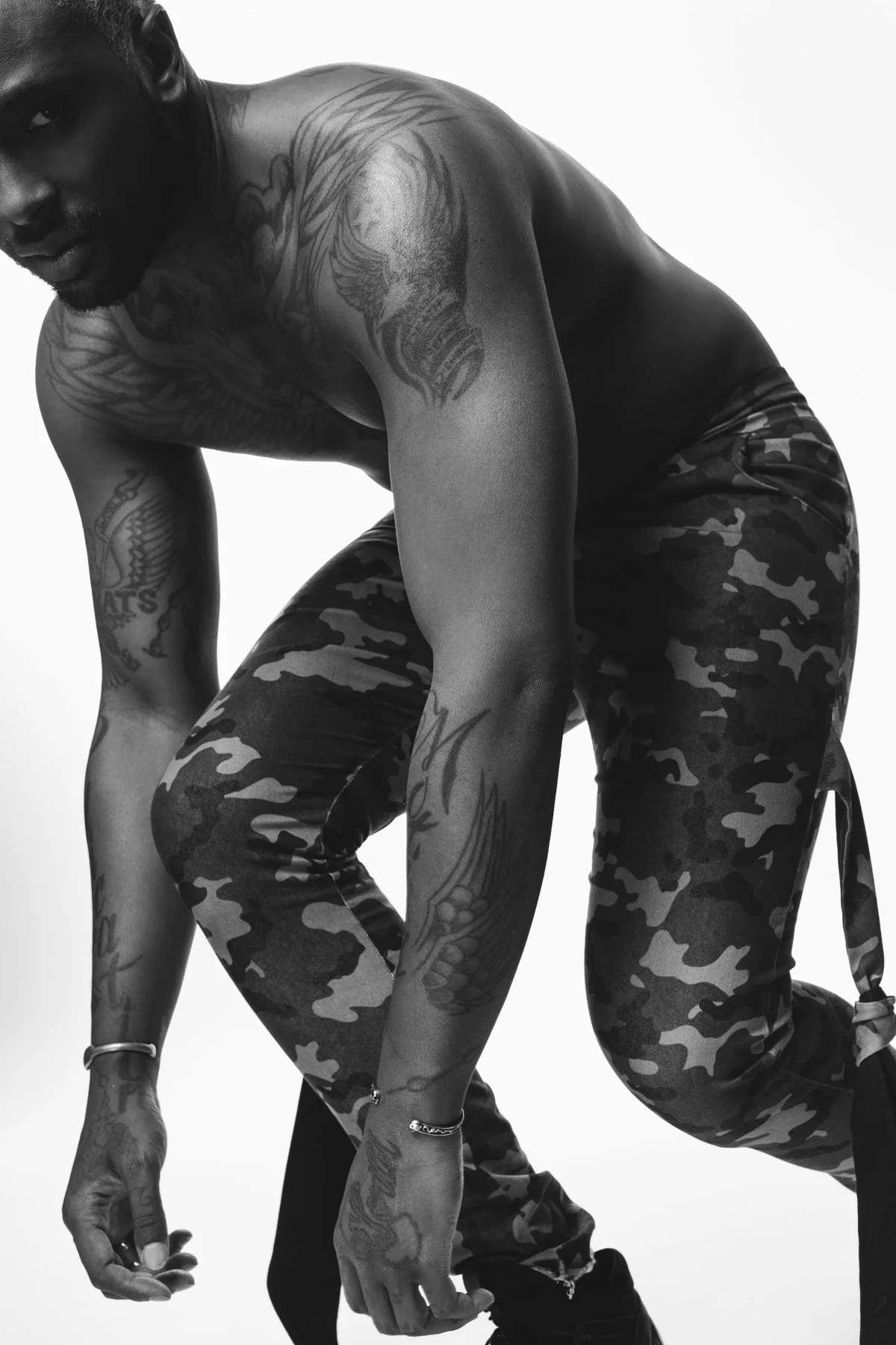 Black and white photo of a shirtless person with tattoos, wearing camo pants.