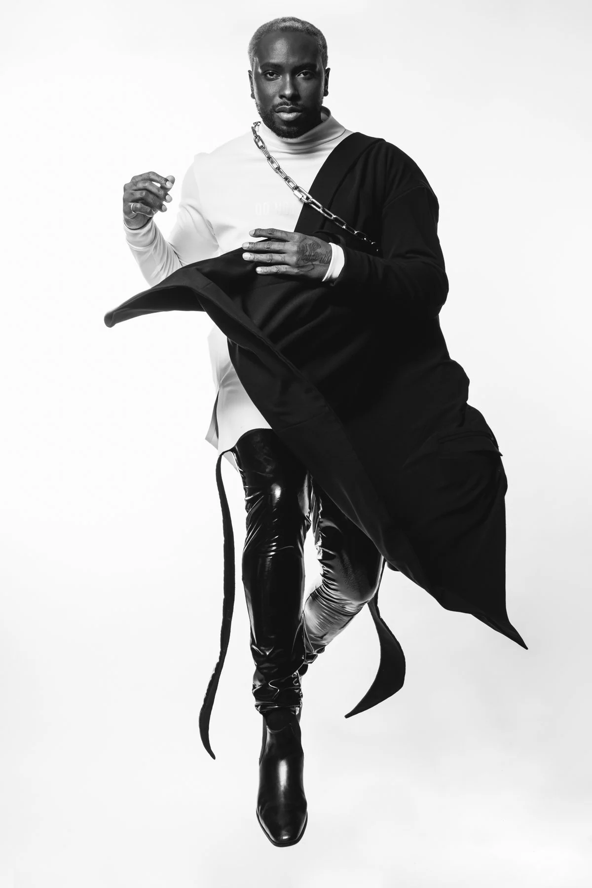 Monochrome photo of a person wearing futuristic clothing, including a white top with a chain strap, black flowing garment, shiny black pants, and boots, in a dynamic pose with a white background.