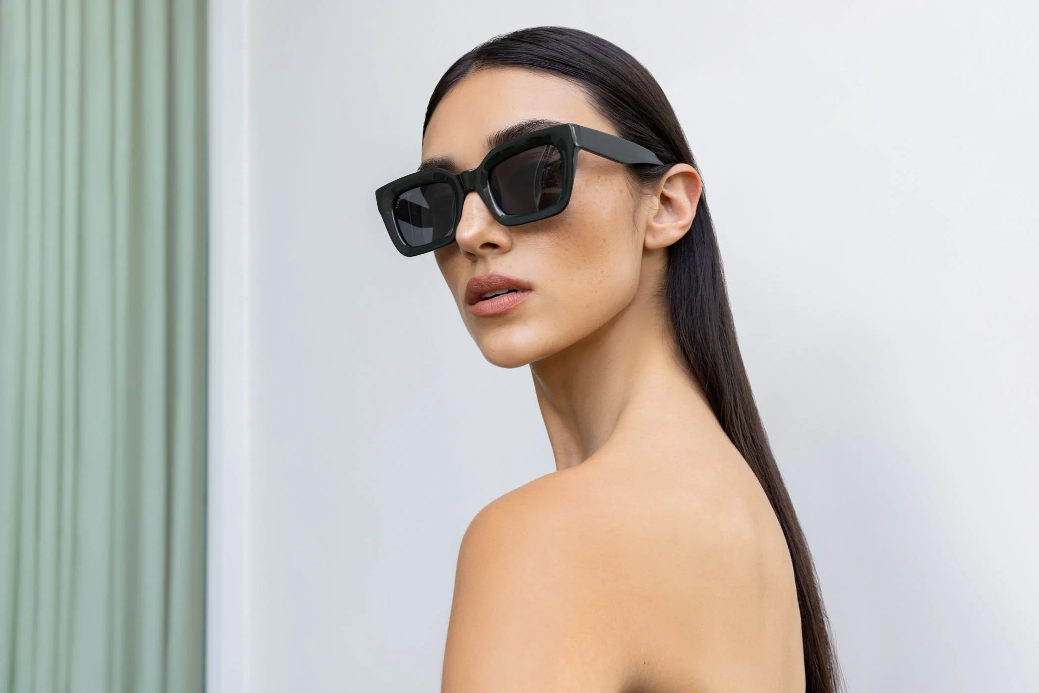 Person wearing black rectangular sunglasses, bare shoulders, with long dark hair, looking over shoulder in front of a neutral background.