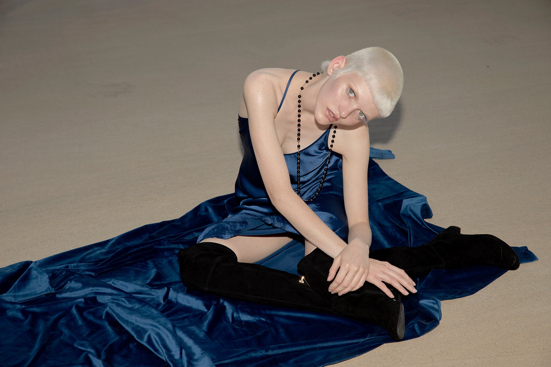 Person sitting on the ground wearing a blue satin dress, black thigh-high boots, and a black beaded necklace.
