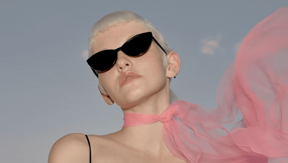Person with platinum blonde hair wearing black sunglasses and a pink sheer fabric around their neck, against a clear sky.