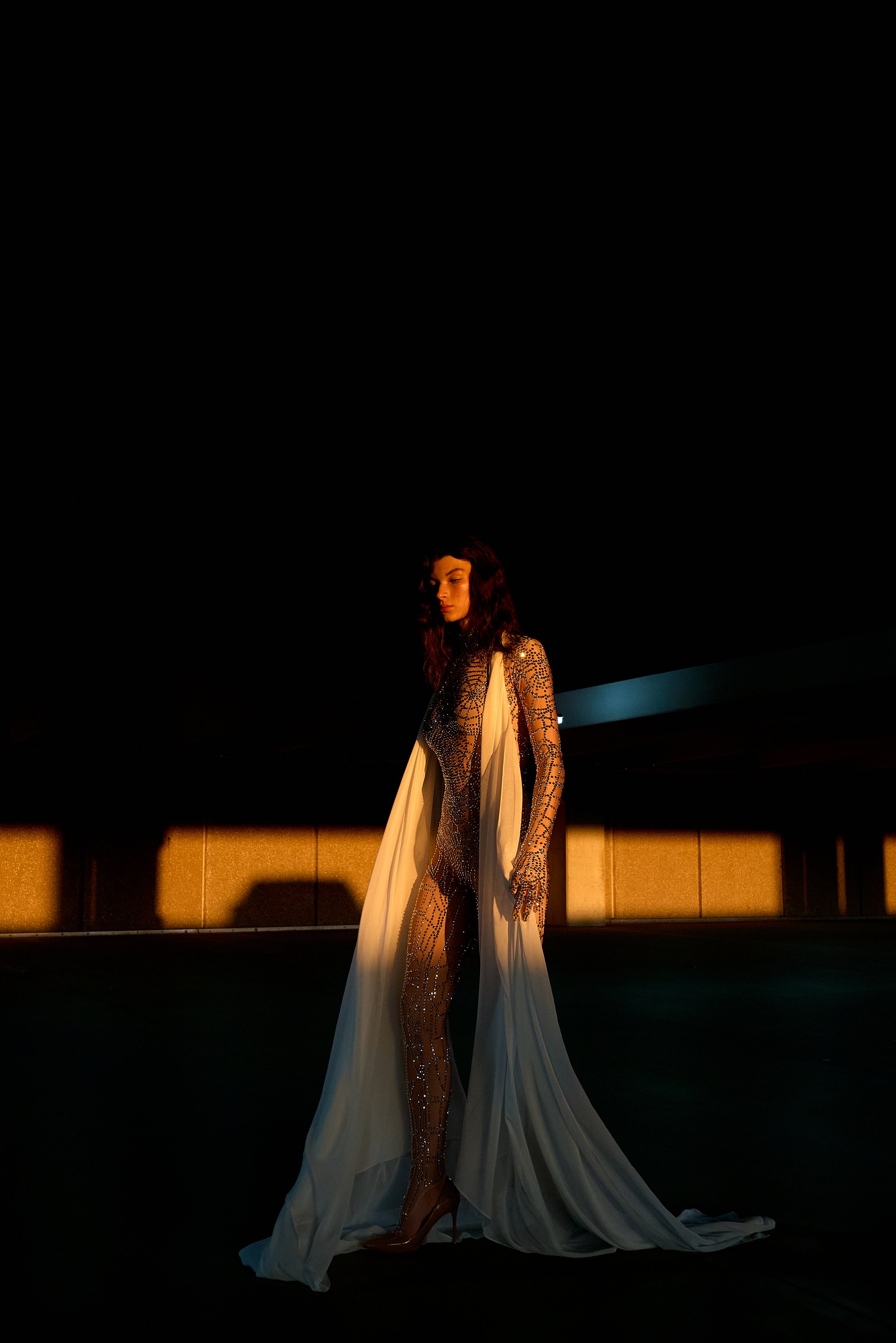 Person wearing a glittering bodysuit with white flowing fabric, standing in dramatic lighting.