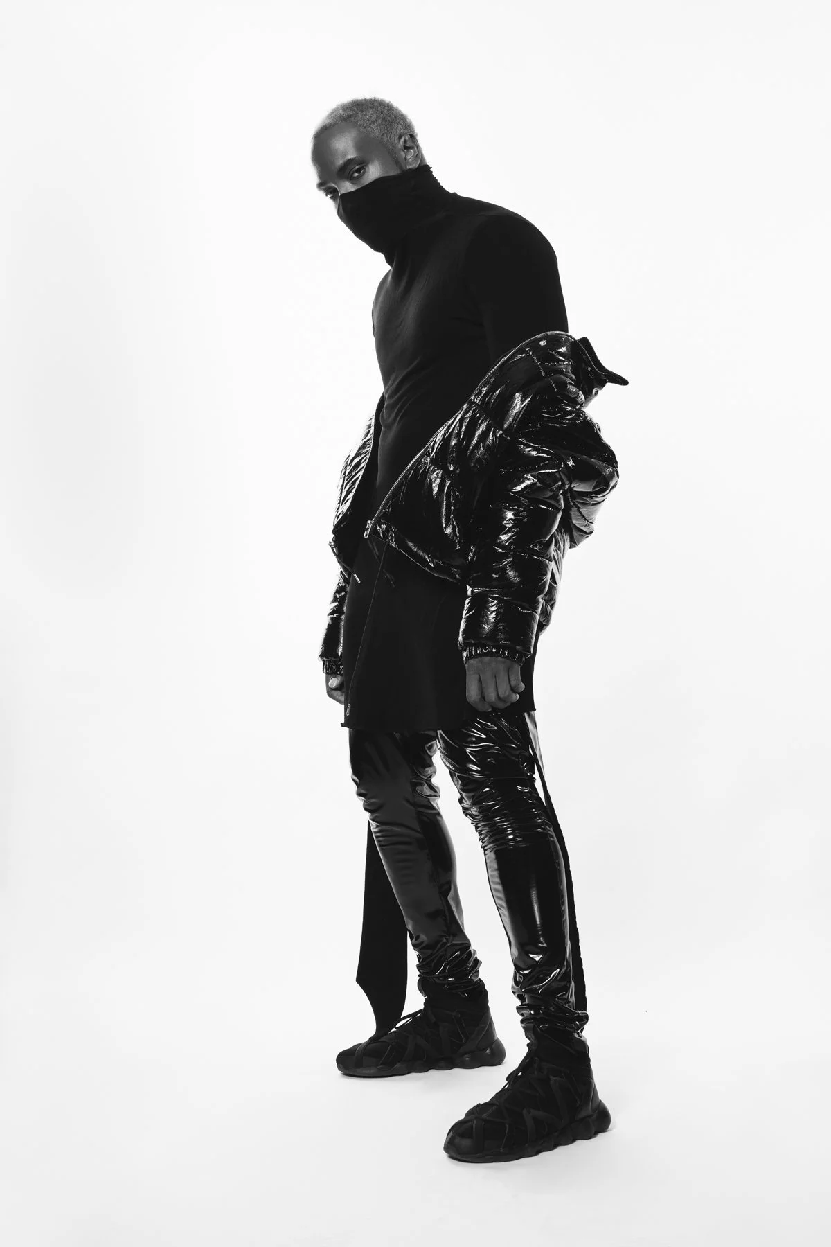 Individual in a black turtleneck and shiny black jacket, wearing dark, glossy pants and sneakers in a monochrome studio setting.