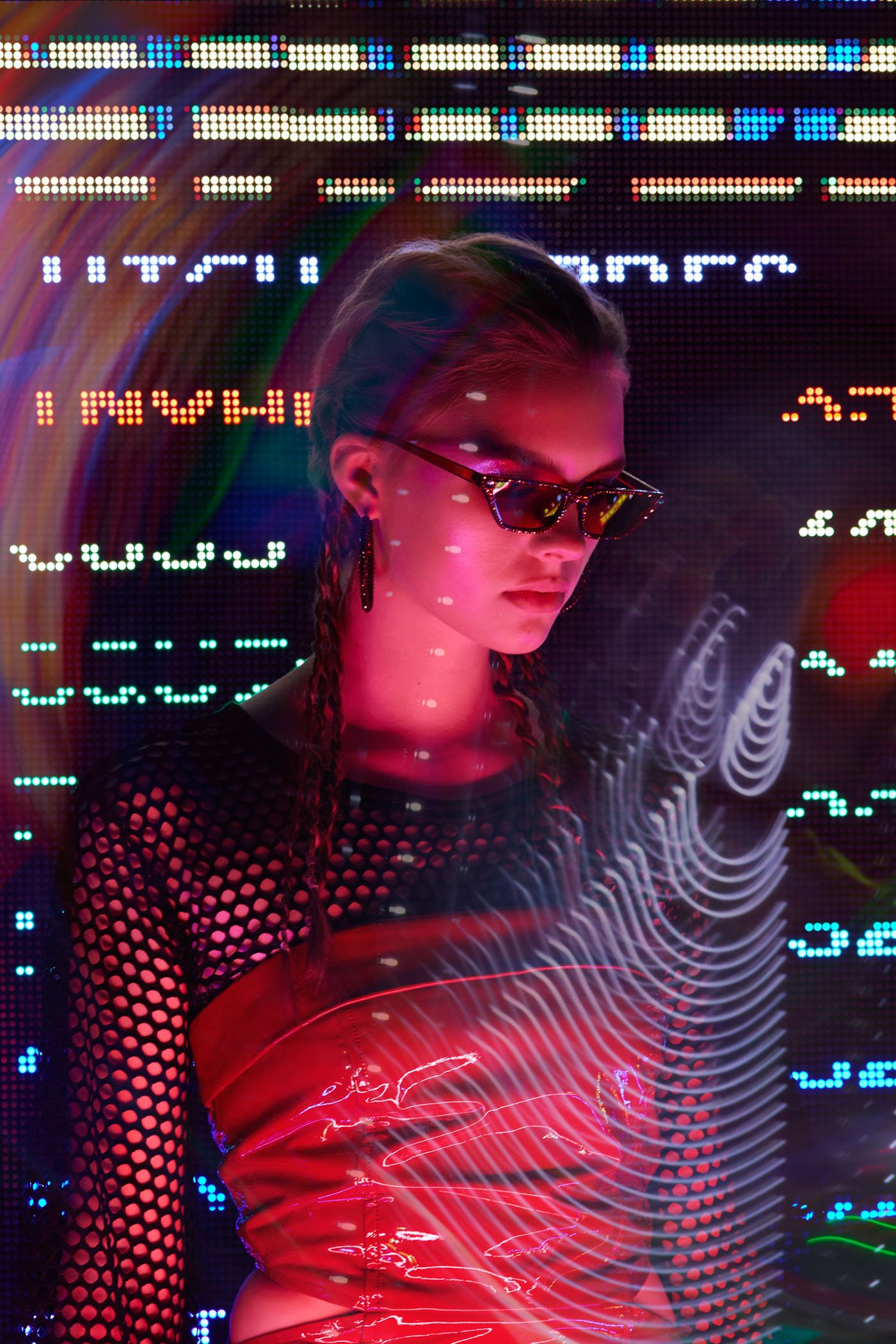 A person wearing sunglasses and a shiny red outfit stands in front of a vibrant digital display featuring colorful, abstract patterns and symbols.