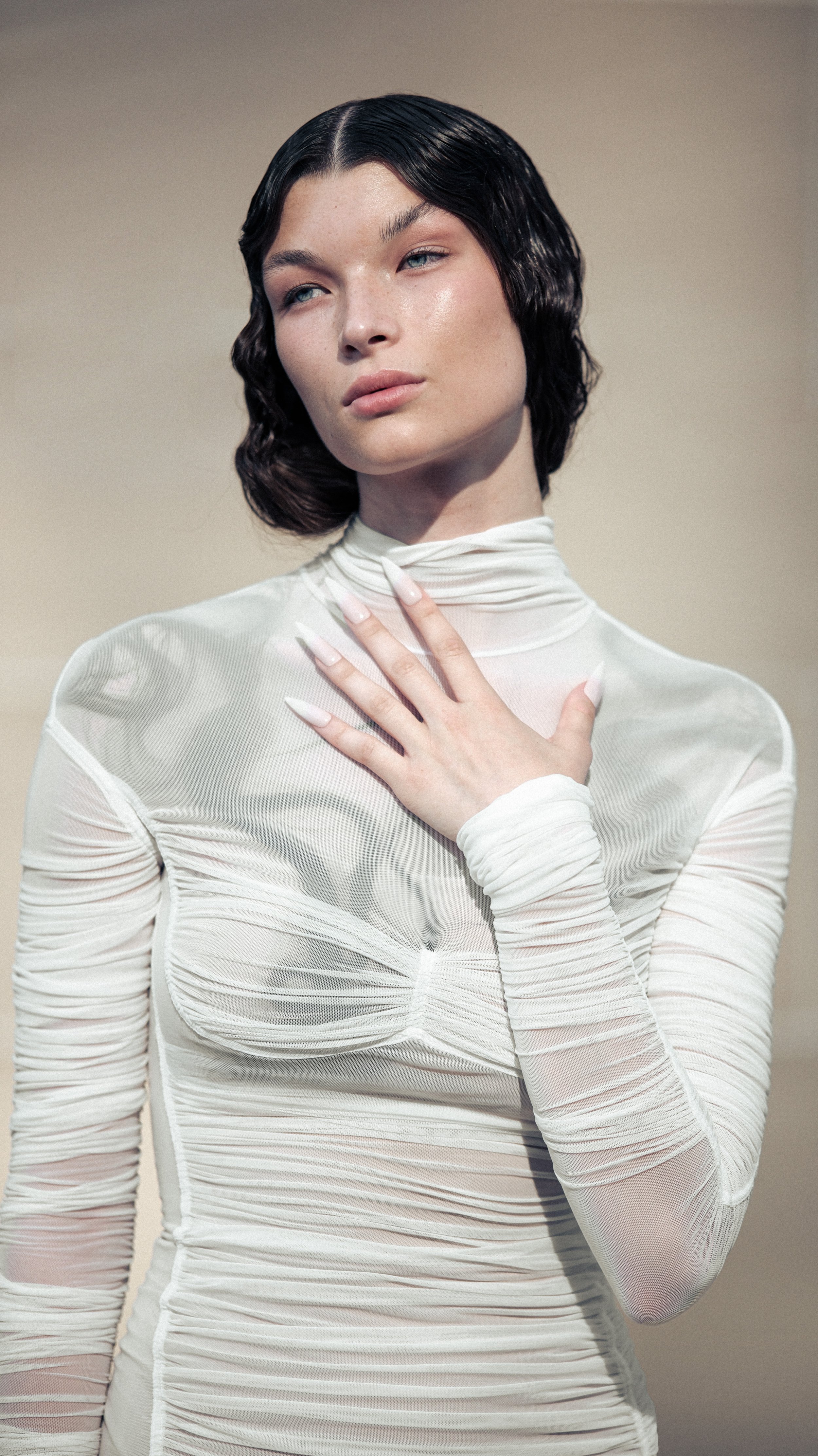 Person wearing a sheer white long-sleeve top with ruched details, styled with side-parted wavy hair and a hand placed on the chest.