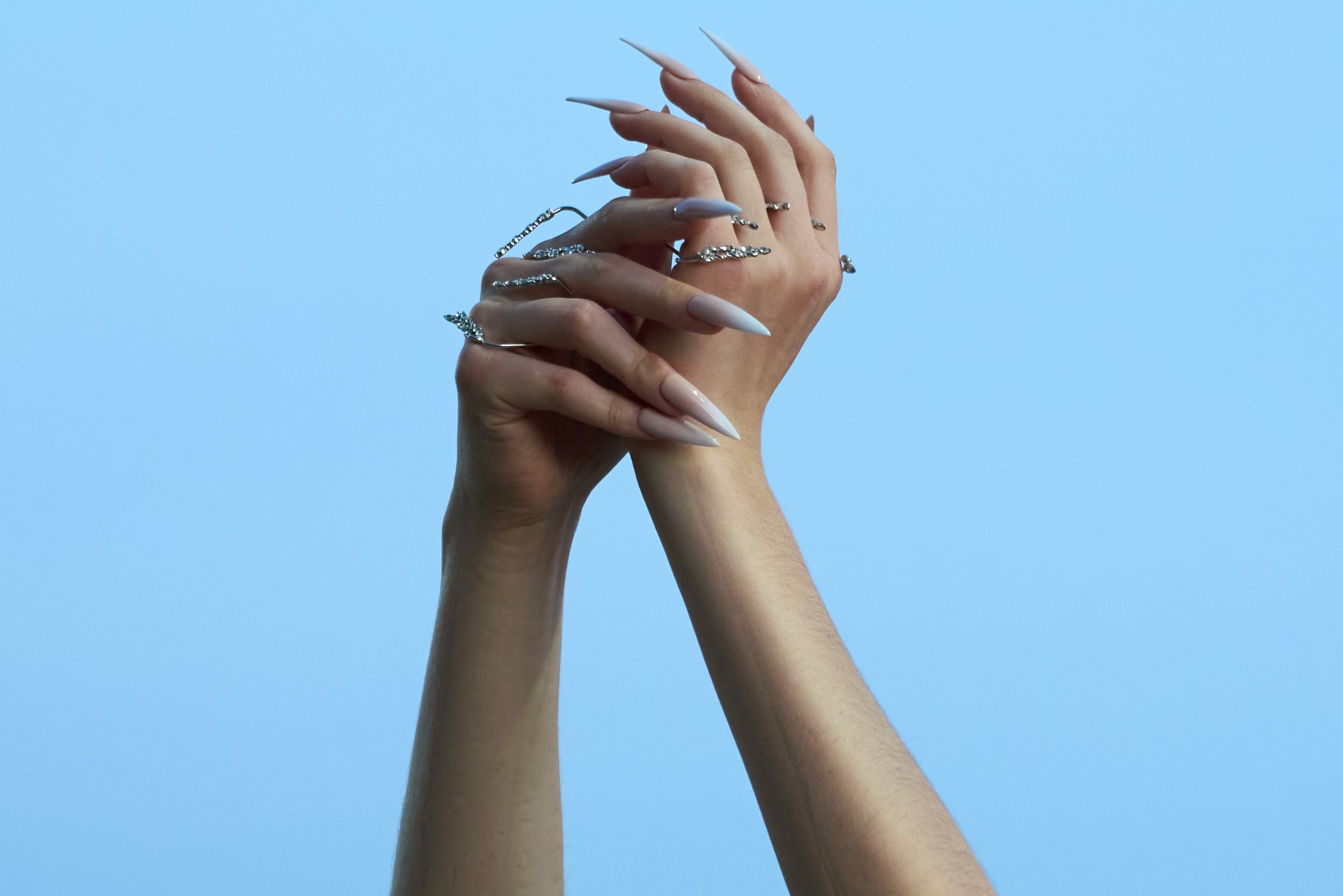 Hands with long, pointed acrylic nails and silver rings against a blue background.