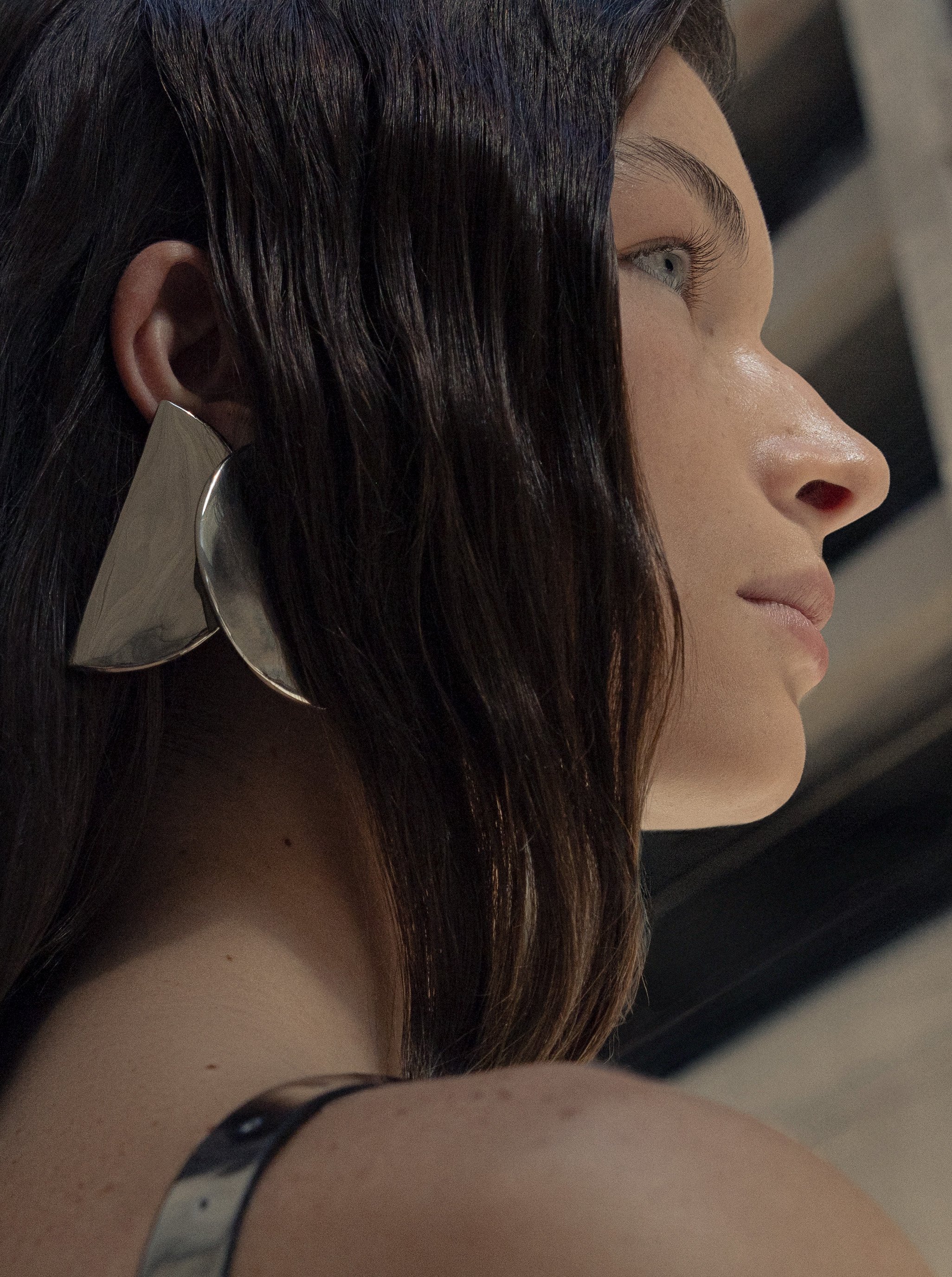 Close-up of a woman's face in profile, with long brown hair and a large, abstract silver earring.