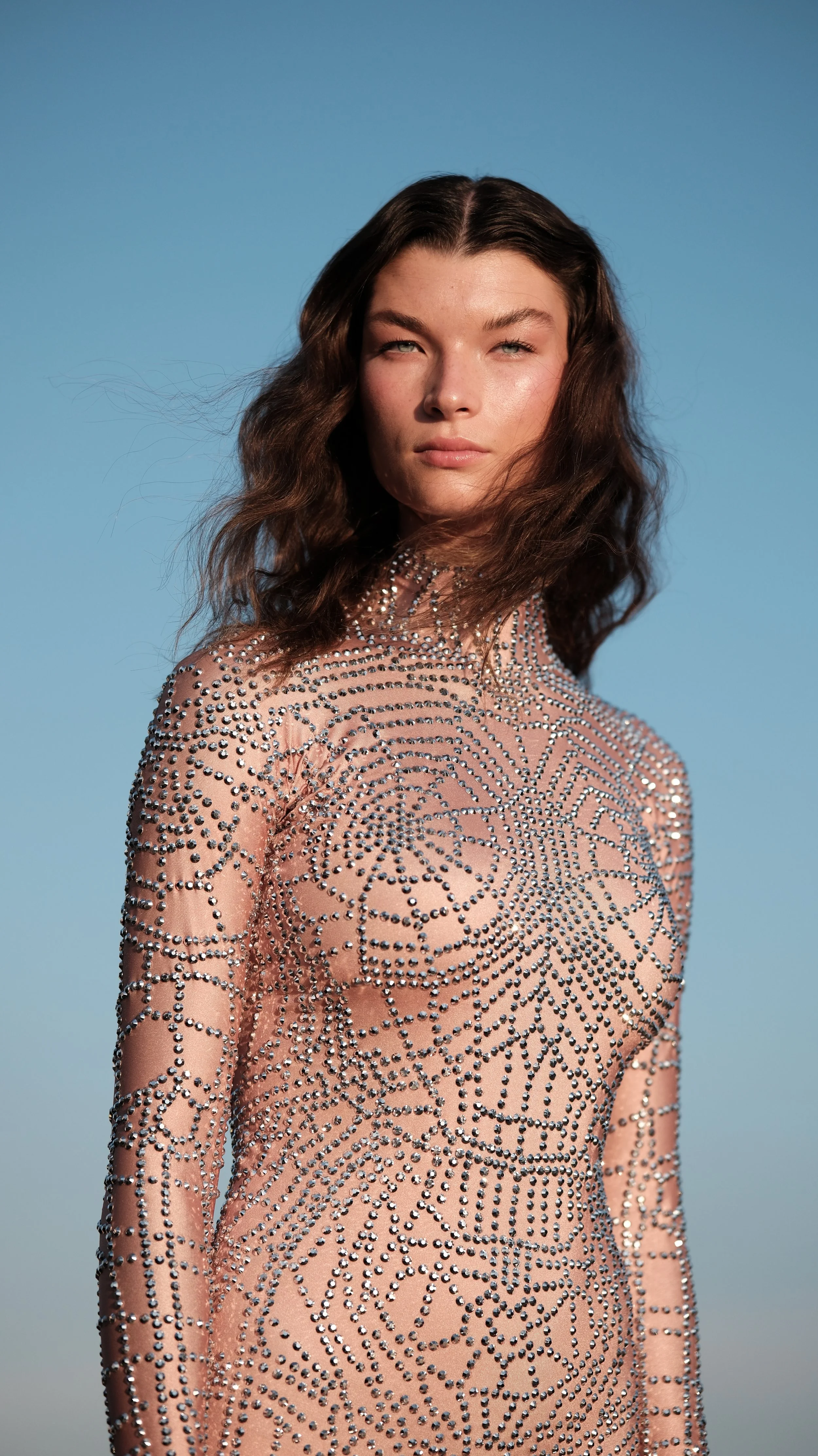 A young woman with wavy brown hair stands outdoors against a clear blue sky, wearing a long-sleeved, form-fitting, nude-colored dress decorated with silver rhinestones in intricate swirling patterns.