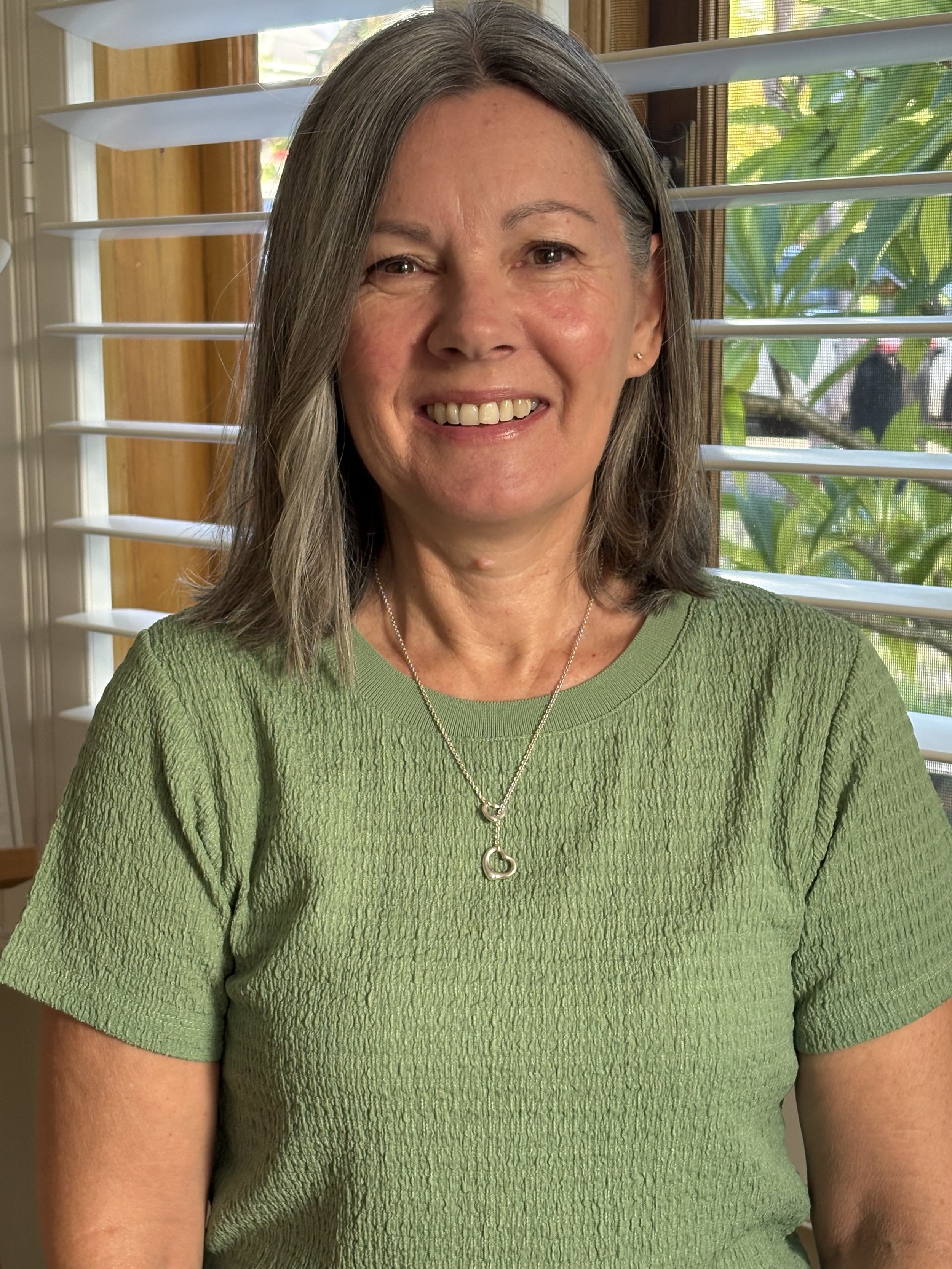 A smiling woman with shoulder-length gray hair wearing a light green textured T-shirt and a silver necklace with a heart-shaped pendant, sitting in front of a window with white blinds and outside greenery visible.