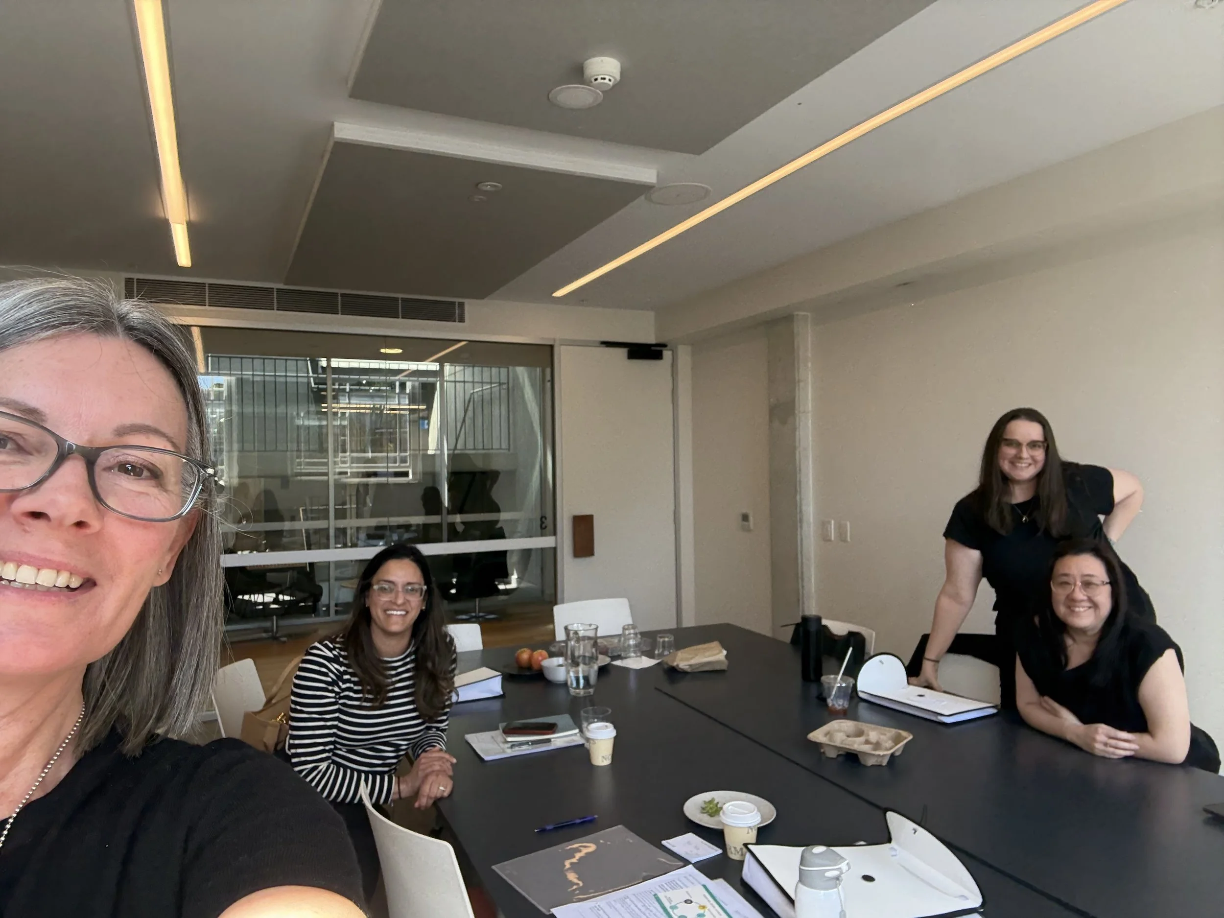 Four women in a conference room, one taking a selfie, with a large meeting table, notebooks, drinks, and a glass wall.