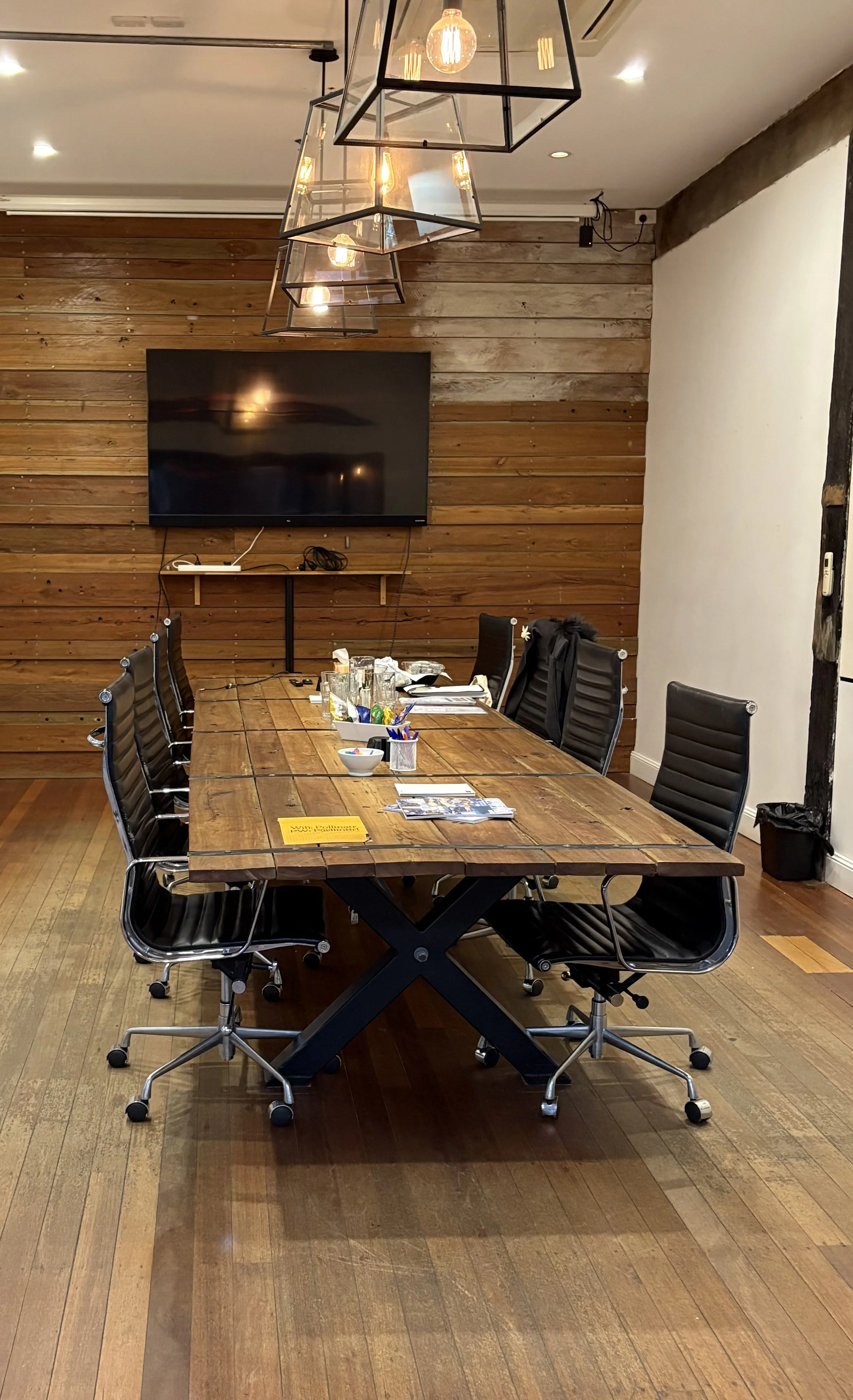 A conference room with a large wooden table surrounded by black leather office chairs, a flat-screen TV mounted on a wooden wall, and modern pendant lights hanging from the ceiling.