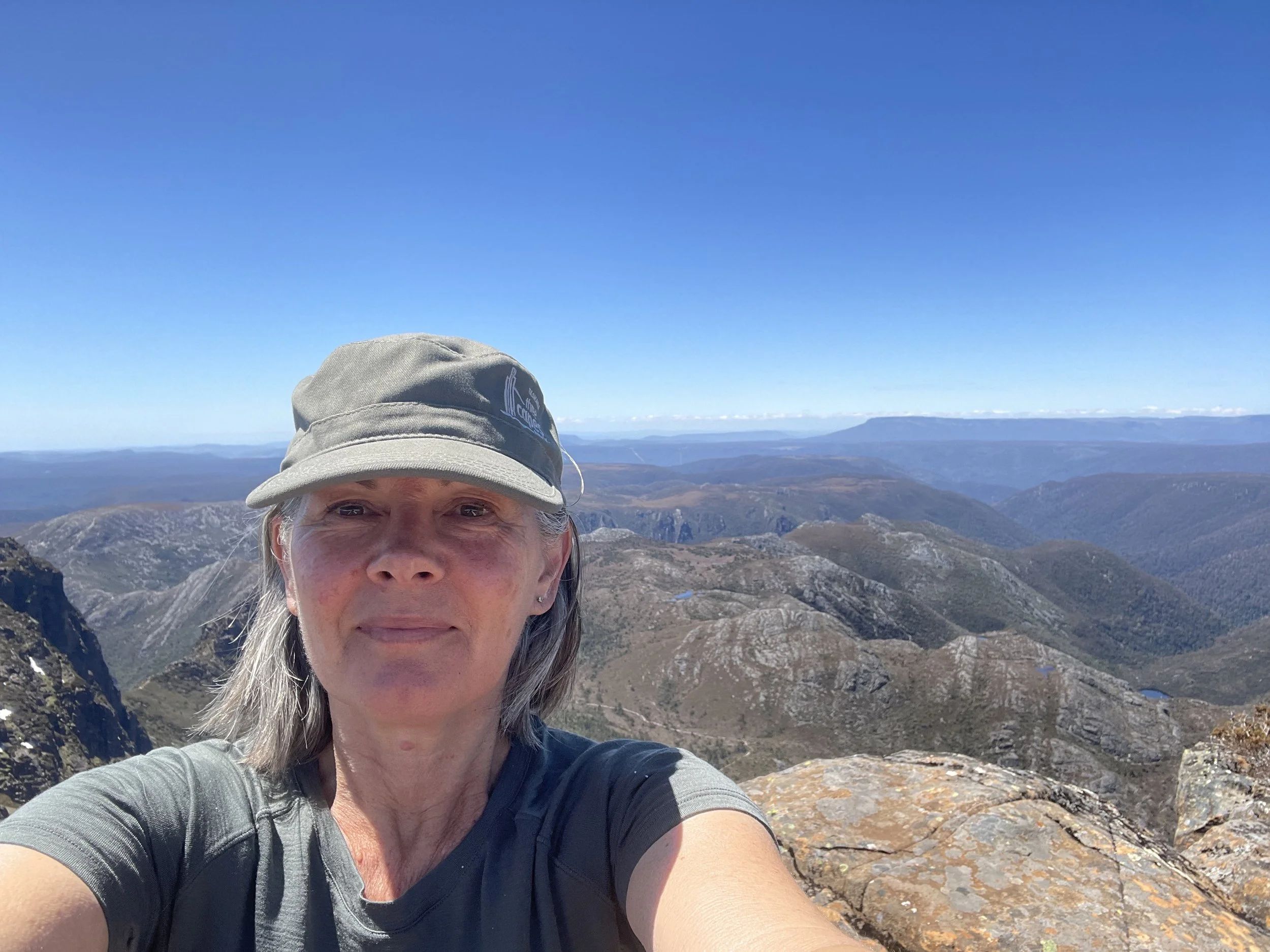 A woman wearing a gray cap and t-shirt taking a selfie at a mountain summit with panoramic mountain views and a clear blue sky in the background.