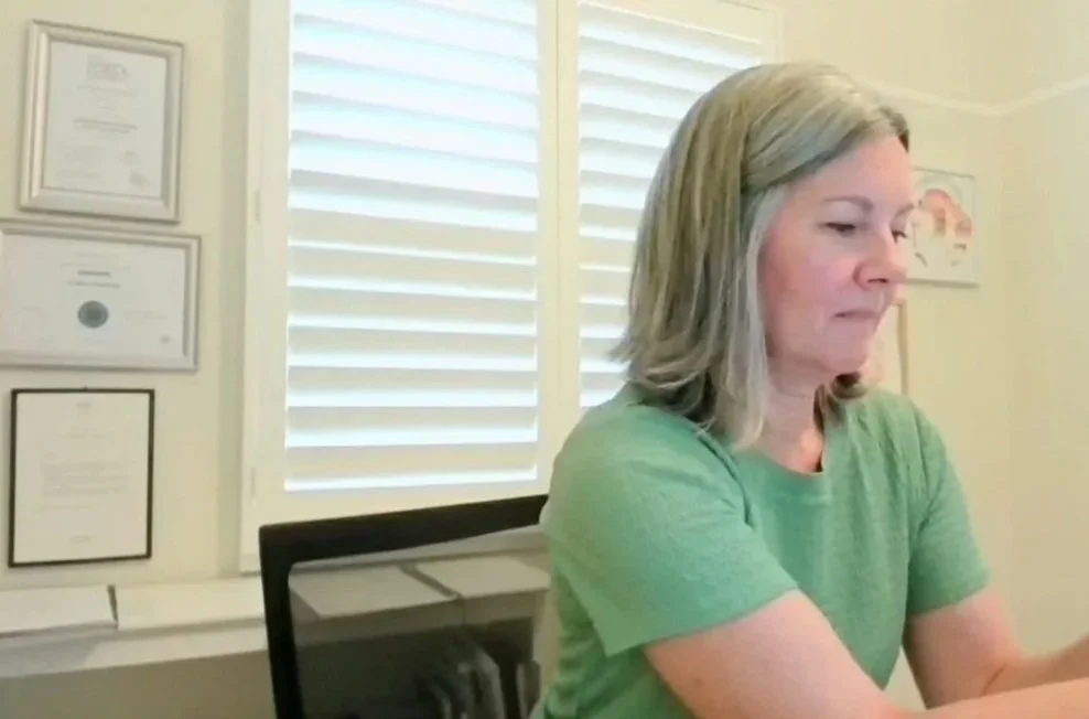 Cathy Hockey coach with gray hair wearing a green shirt sitting at a desk looking down at her phone in a room with framed certificates on the wall and a window with shutters.