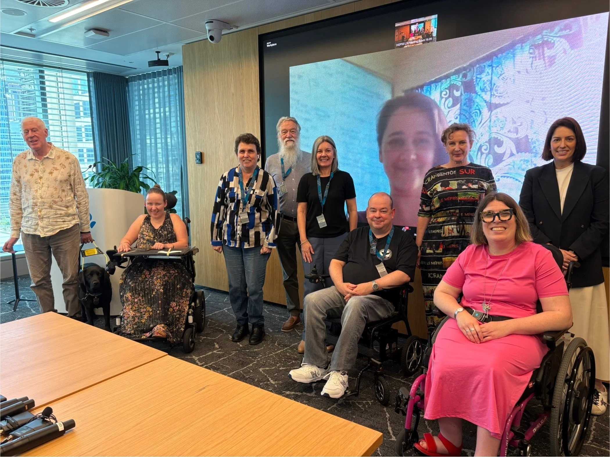 Group of diverse individuals, some in wheelchairs, gathered in a conference room for a photo. There are nine people, including a young girl in a wheelchair with a guide dog and others smiling, standing or seated. A large screen shows a video call with a woman’s face, and the room has large windows with cityscape views.