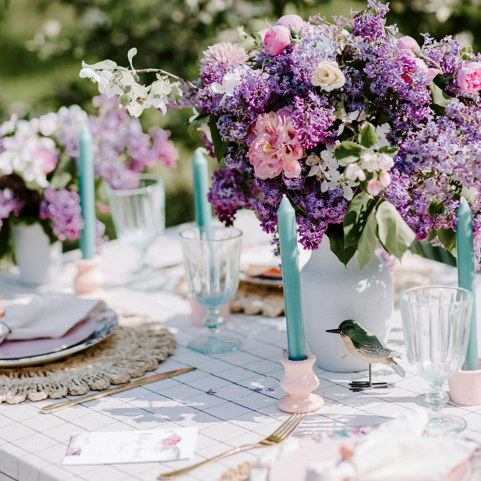 A beautifully set outdoor dining table decorated with a large floral centerpiece featuring purple, pink, and white flowers, white candle holders with pink bases and light blue candles, glassware, and pastel-colored details.