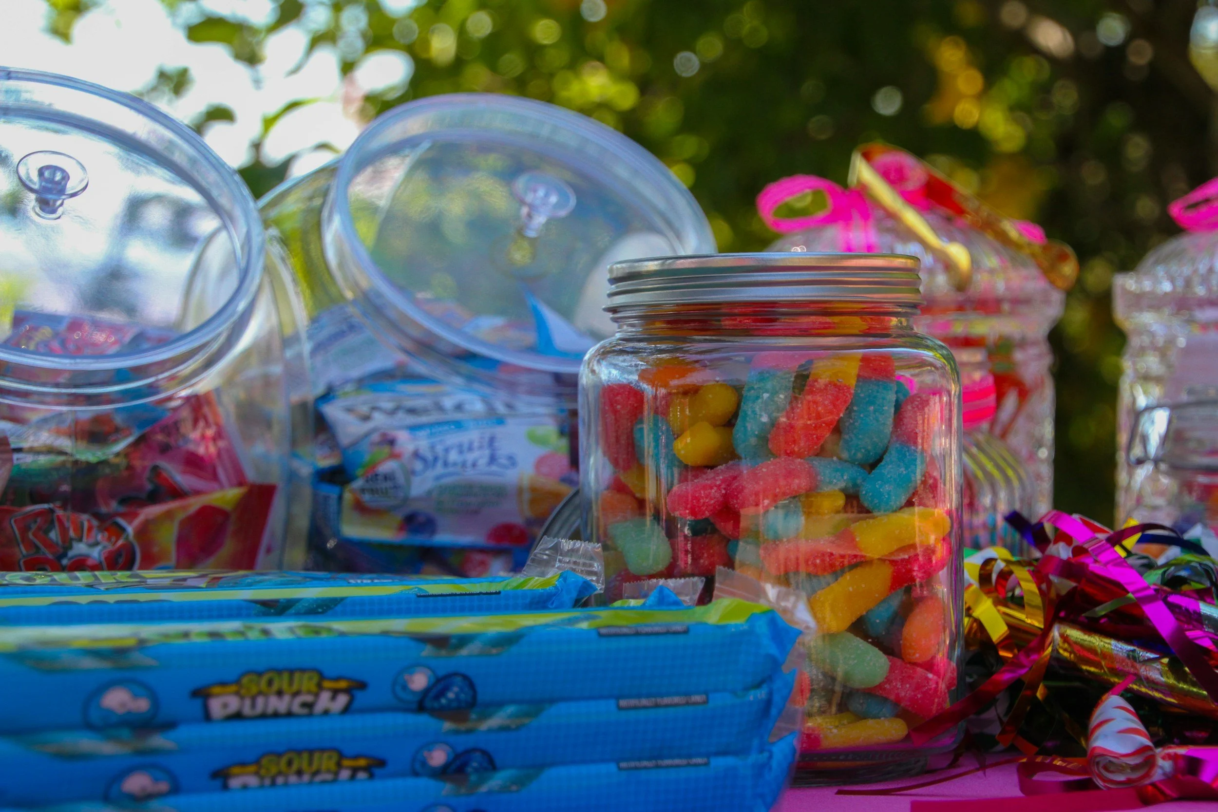 An assortment of candies in jars and packaging, including gummy worms, sour punch strips, and other assorted sweets, on a colorful table with a blurred outdoor background.