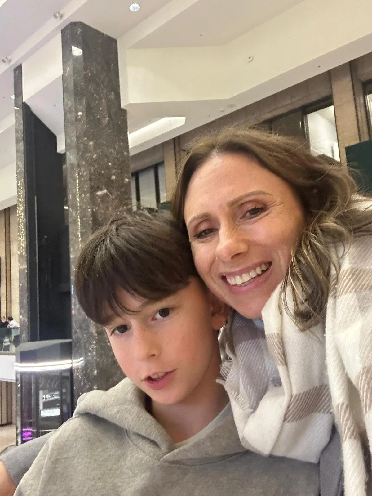 A woman and a young boy taking a selfie together in an indoor setting, smiling with their heads close. The woman has shoulder-length wavy brown hair and is wearing a light-colored jacket. The boy has straight brown hair and is wearing a gray hoodie.