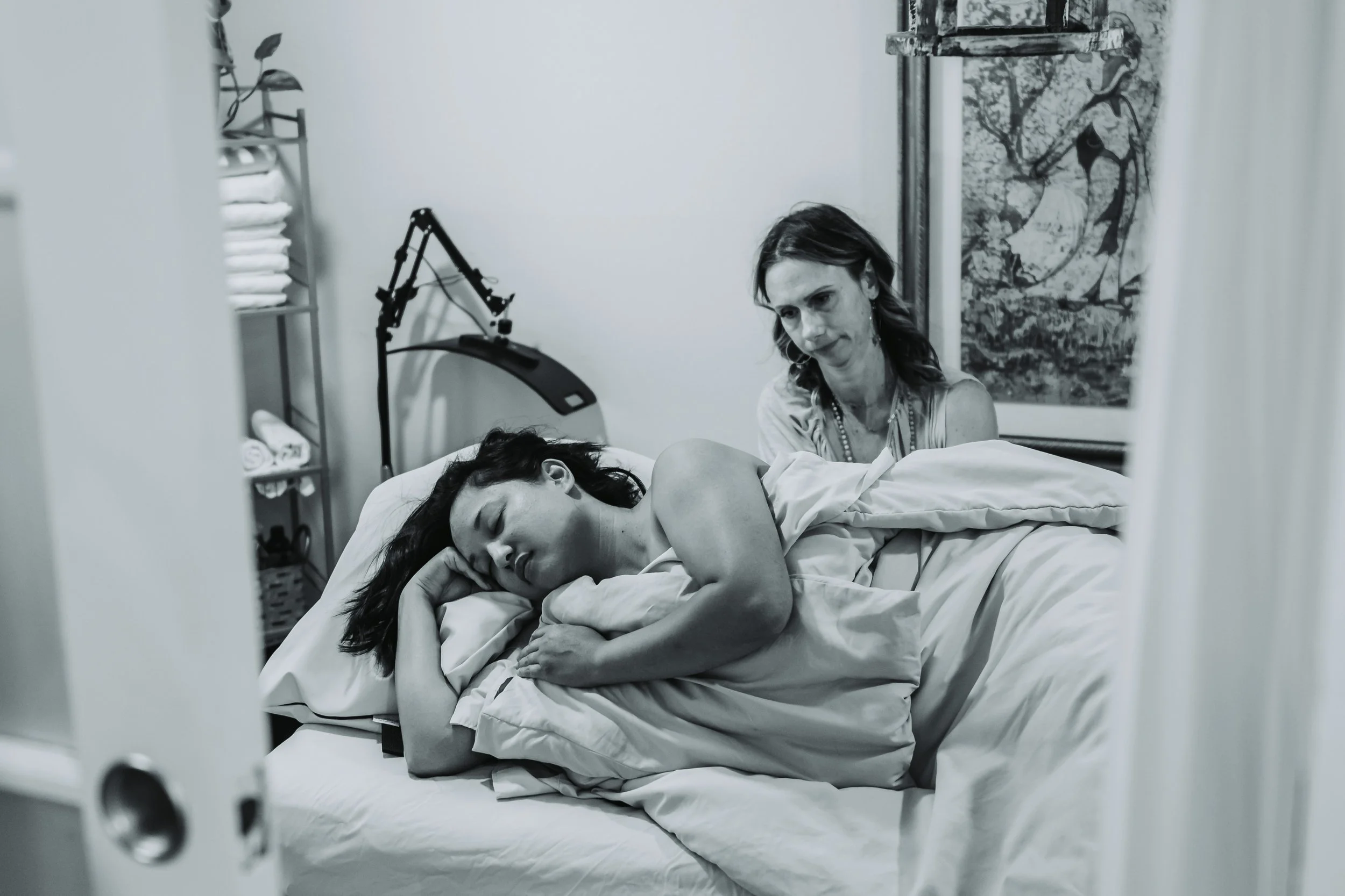A woman lying in a hospital bed with her eyes closed, resting her head on her hand, while another woman sits beside her, looking concerned. The hospital room has medical equipment, shelves with supplies, and a decorative wall hanging.