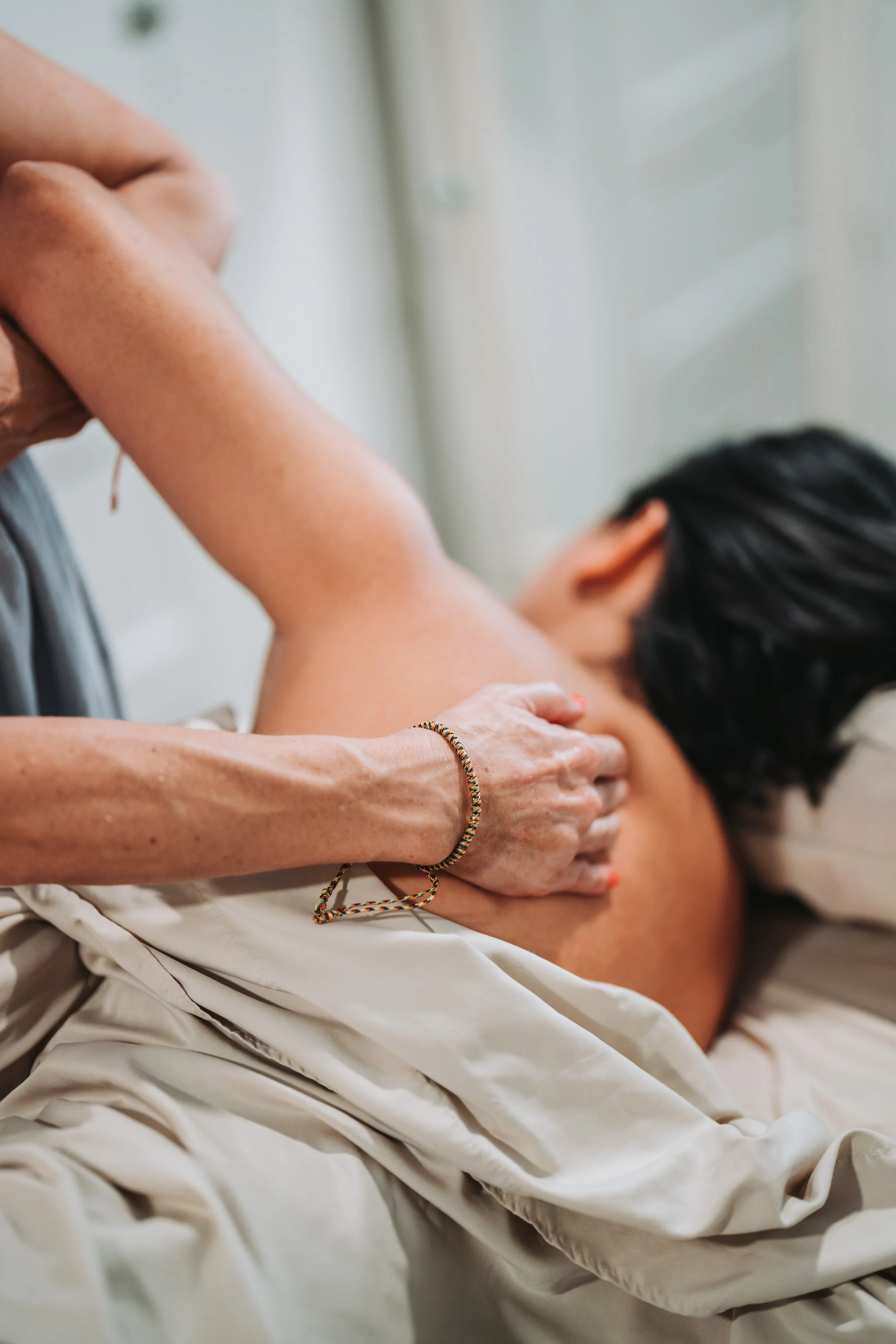 A person receiving a massage or physical therapy on their back, lying face down on a bed or table with a white sheet draped over lower body, with the practitioner using their hands to massage or manipulate the person's back.