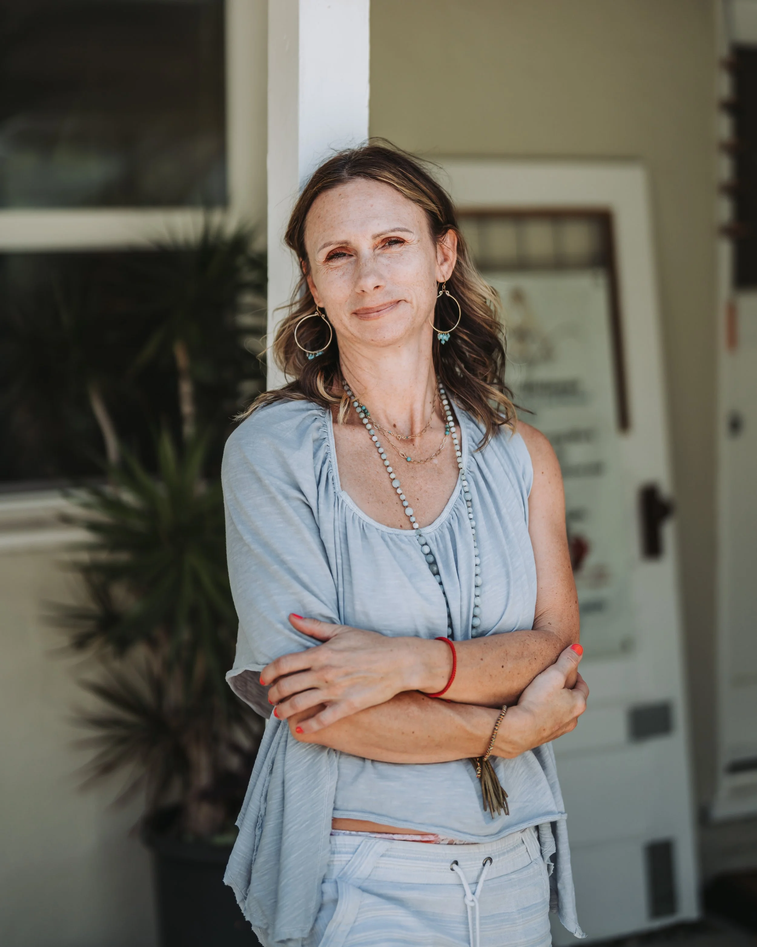 A woman with brown hair and freckles stands outdoors near a building, crossing her arms and smiling gently. She is wearing a light blue top, striped shorts, and jewelry including earrings and necklaces.