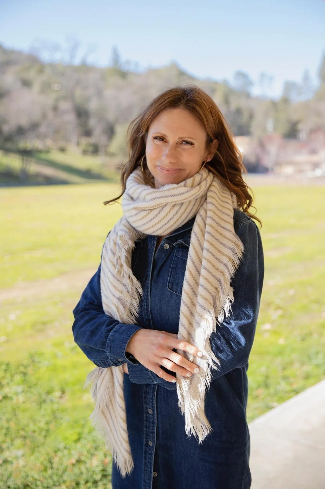 A woman with red hair standing outdoors on a sunny day, wearing a dark denim jacket and a light striped scarf, with a grassy park and trees in the background.