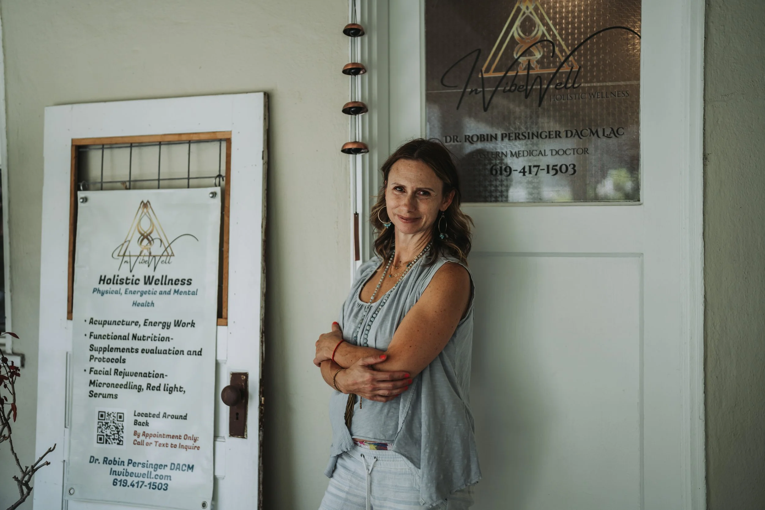 A woman standing with her arms crossed, smiling in front of a signage for a holistic wellness clinic. The sign includes the name Dr. Robin Persinger DACM, and a list of services offered, such as acupuncture, energy work, functional nutrition, facials, microneedling, and serums. The woman wears a light-colored sleeveless top with jewelry.