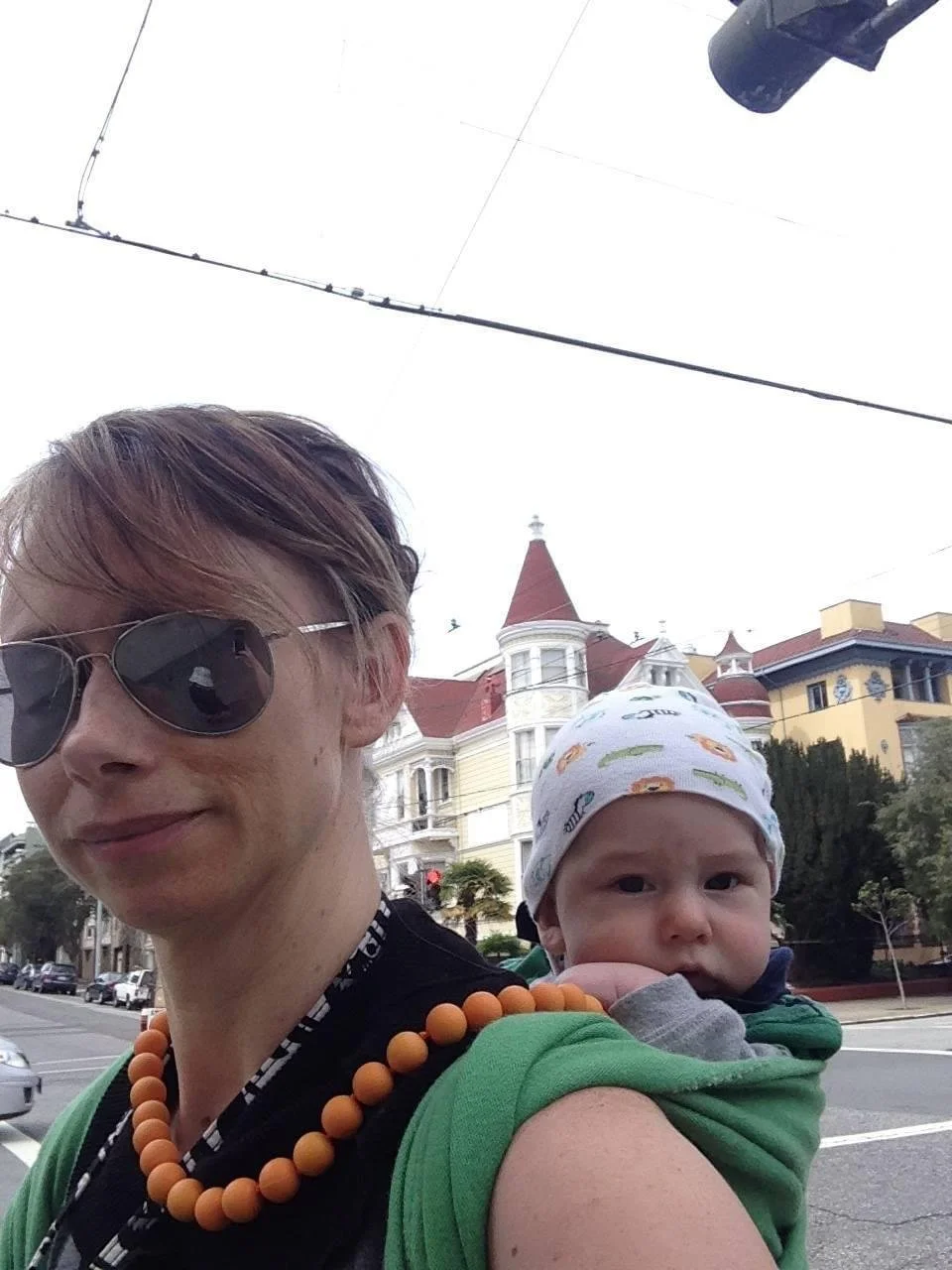 A woman wearing sunglasses and a beaded necklace carries a child on her shoulder. The woman is smiling slightly, and the child is looking directly at the camera with a neutral expression. Both are outdoors in front of a row of Victorian-style houses on a city street.
