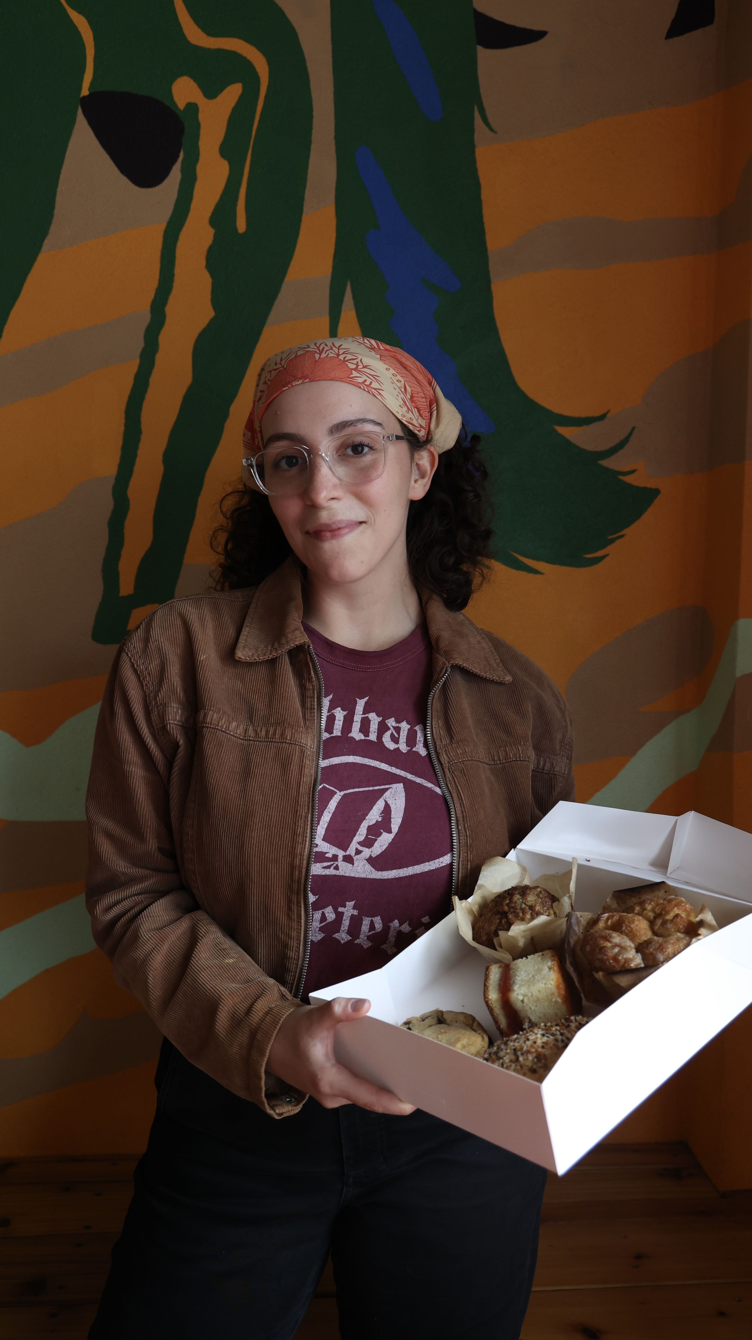 Woman holding a box of assorted baked goods, standing in front of a colorful abstract mural.