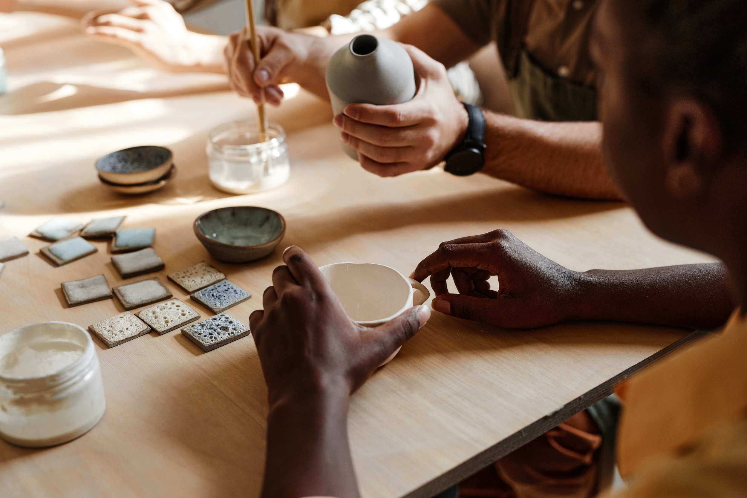 People creating pottery, working with clay and glazes at a table with various ceramic tiles and bowls.
