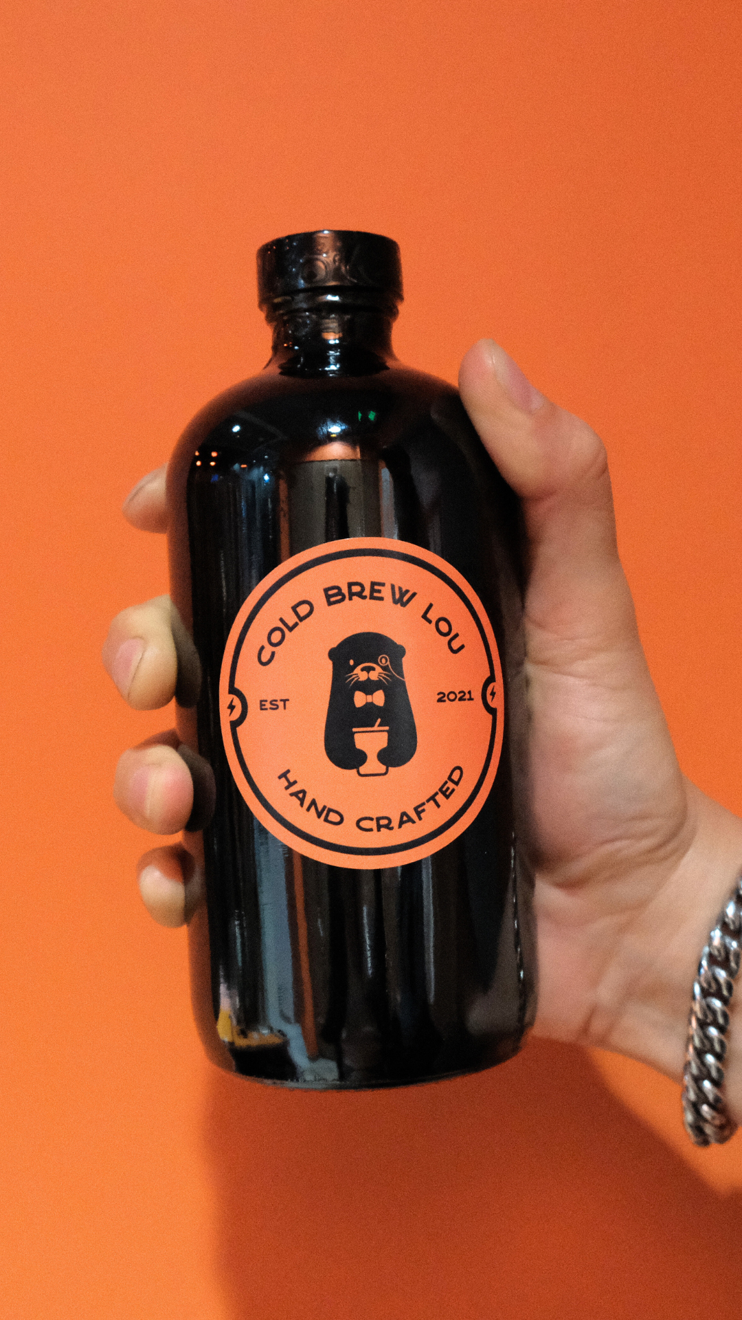 A hand holding a black bottle with a round orange label that reads "Cold Brew Lou" and features a bear with a bowtie holding a coffee cup, against an orange background.