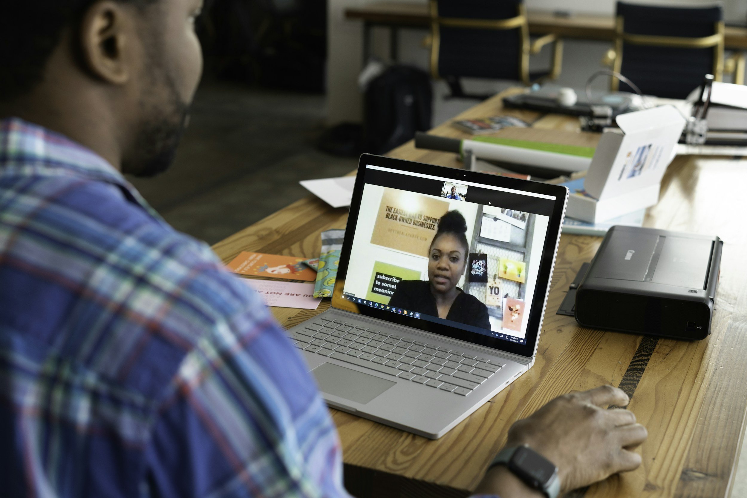 Person sitting at a desk participating in a video call with a woman on a laptop, surrounded by office supplies and papers.