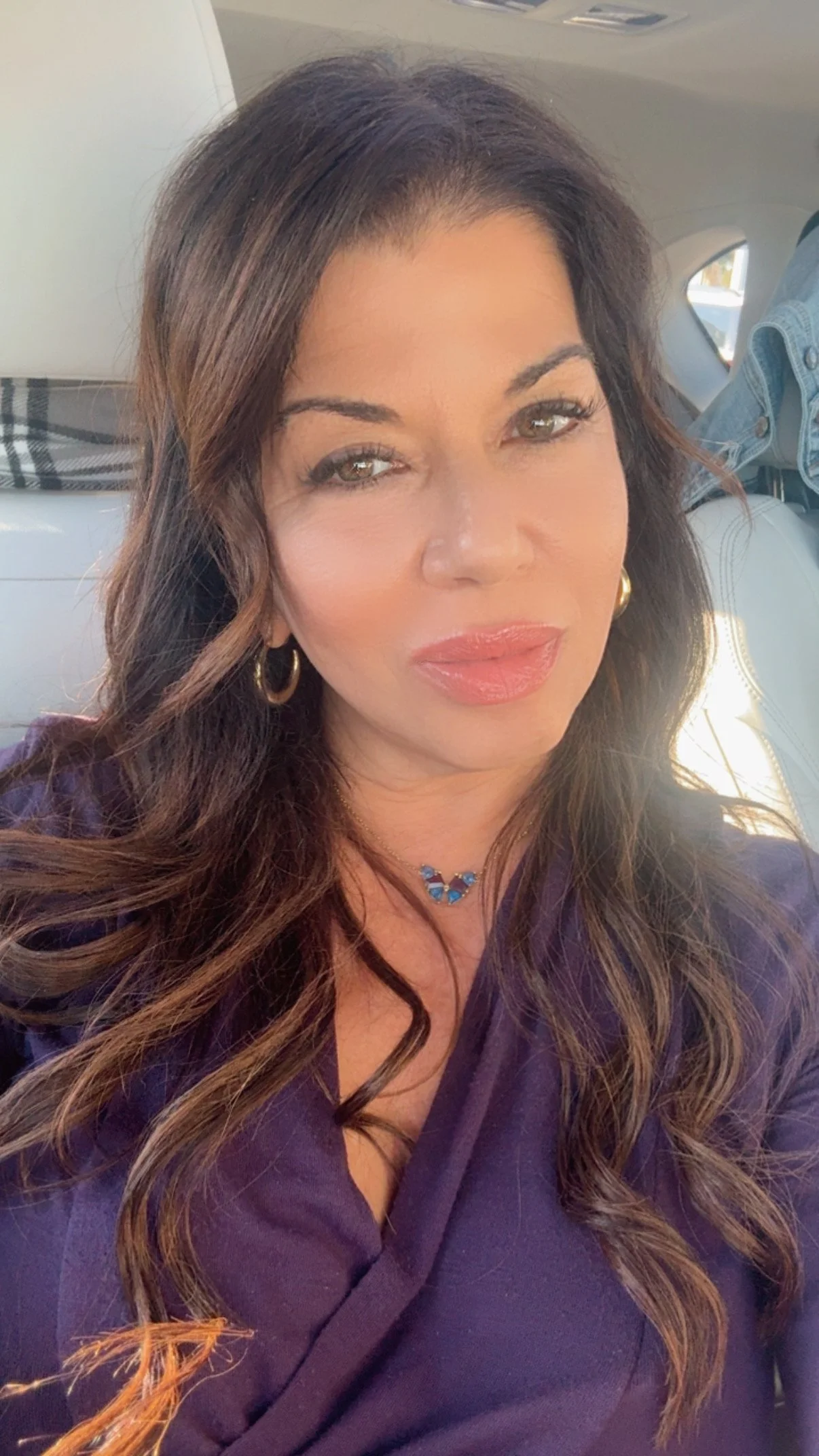 A woman with long, wavy brown hair taking a selfie inside a car. She is wearing gold hoop earrings, a blue necklace, and a purple top. Sunlight is shining on her face.